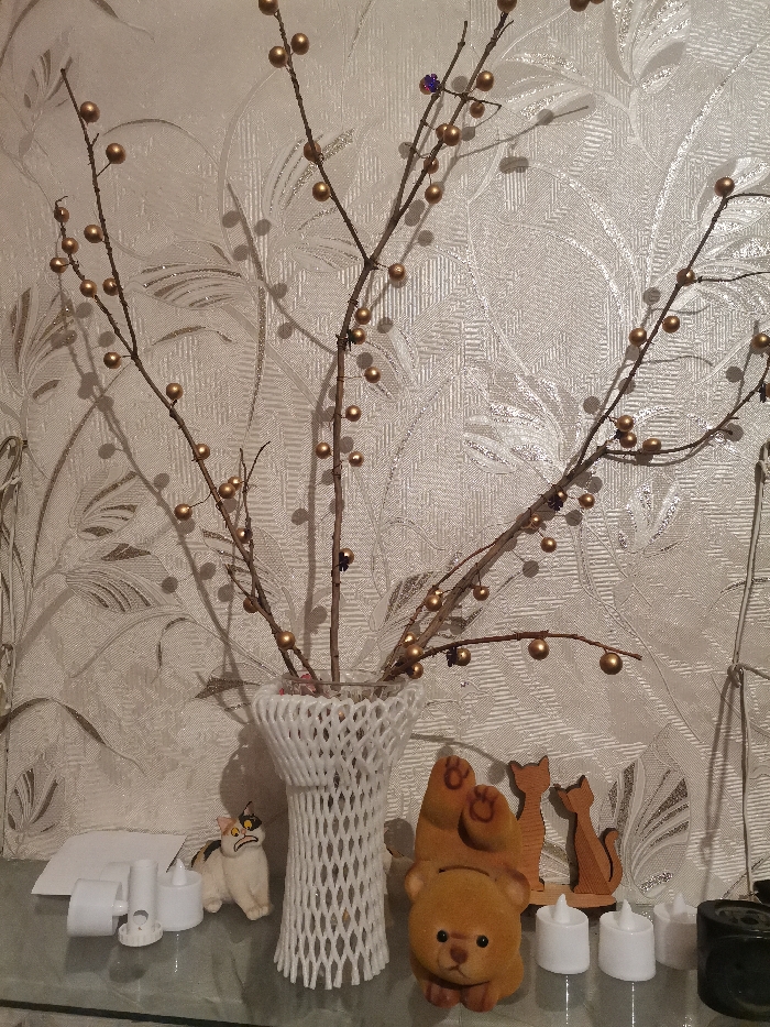 This image captures a close-up indoor scene featuring a decorative arrangement on a clear, reflective surface, set against a light-colored, textured wallpaper.
At the center, a vase holds several bare branches. The vase itself is covered with a white, open-weave, mesh-like material, giving it a distinct patterned appearance. The branches are adorned with numerous small, spherical, golden-colored beads, resembling berries or pearls, giving them a festive and ornamental quality. A few small, darker, possibly purple or brown, decorative elements are also scattered among the golden beads on the branches. The branches cast prominent, slightly blurred shadows on the wallpaper behind them.
The wallpaper features a subtle, repeating embossed or textured pattern of swirling leaves and floral motifs in a light, possibly off-white or cream hue.
Arranged on the clear, reflective surface in front of the vase are several small decorative items. From left to right:
* Two small, white, cylindrical objects resembling flameless LED candles, accompanied by a small white card or folded paper.
* A small ceramic figurine of a cat with white fur, black patches, and orange markings on its face, depicted with an expression that appears grumpy or startled.
* To the right of the central vase, there is a fuzzy, light brown teddy bear figurine, possibly a coin bank, lying on its back with its paws pointing upwards.
* Behind the teddy bear are two wooden silhouette figurines of cats, one slightly taller than the other, standing upright on a small base.
* Further to the right, three more small, white, cylindrical objects, identical to the ones on the far left, are visible.
* On the far right edge of the frame, a dark, cylindrical object (possibly a container or holder) is partially visible, and a white wire or cord hangs down the side, partially obscured by the branches.
The objects are placed on a transparent surface, likely glass, which shows faint reflections of the items and the light above. The overall lighting suggests an indoor setting, possibly with artificial illumination, creating a warm and decorative ambiance. There is no visible text in the image.