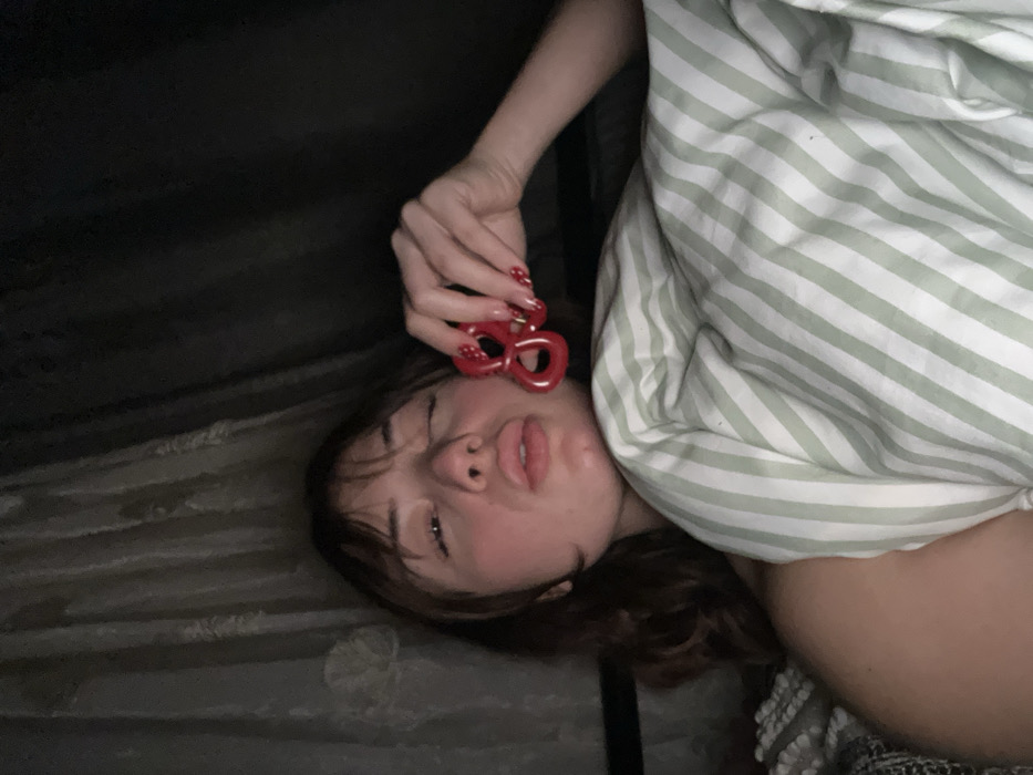 A young woman lies in bed, partly covered by a striped green and white sheet. She holds a pair of red heart-shaped sunglasses above her face with a hand displaying matching red and white nail art. The bed appears dark, and the room is dimly lit, creating a shadowy atmosphere around her. Her expression is serene, eyes partially closed.