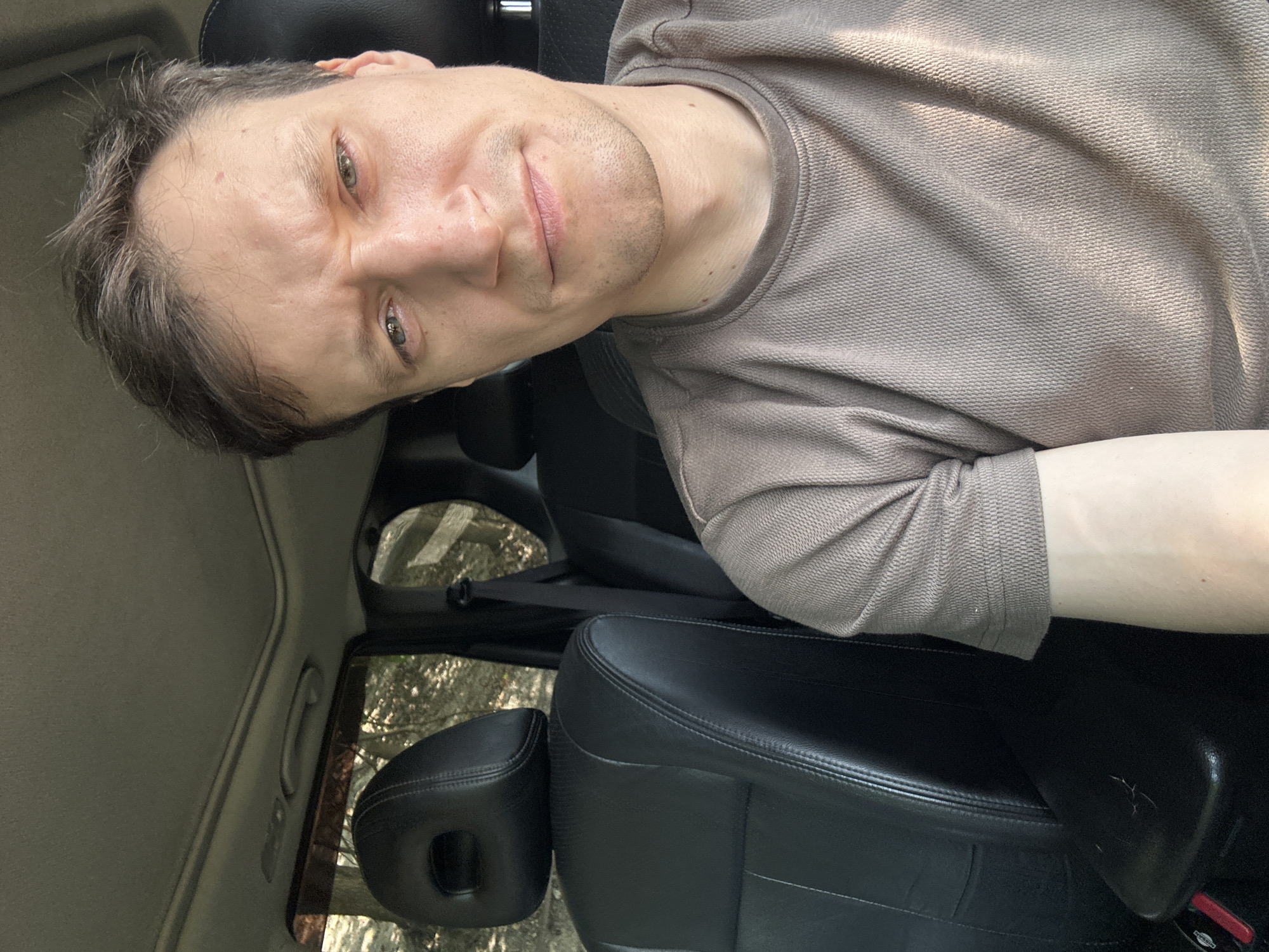 A close-up, waist-up selfie is taken from the perspective of the driver's seat of a vehicle. The man in the image has light skin, short brown hair, and appears to be in his early to mid-thirties. He is wearing a light brown, textured t-shirt and has a slight frown with his eyebrows furrowed. The interior of the car is visible, including a black leather headrest and seat on the left side, and the man's arm resting on the center console on the right. The background is blurred, but appears to be a wooded area with sunlight filtering through the trees, suggesting the vehicle is outdoors, potentially on a rural road near Makiv, Ukraine.
