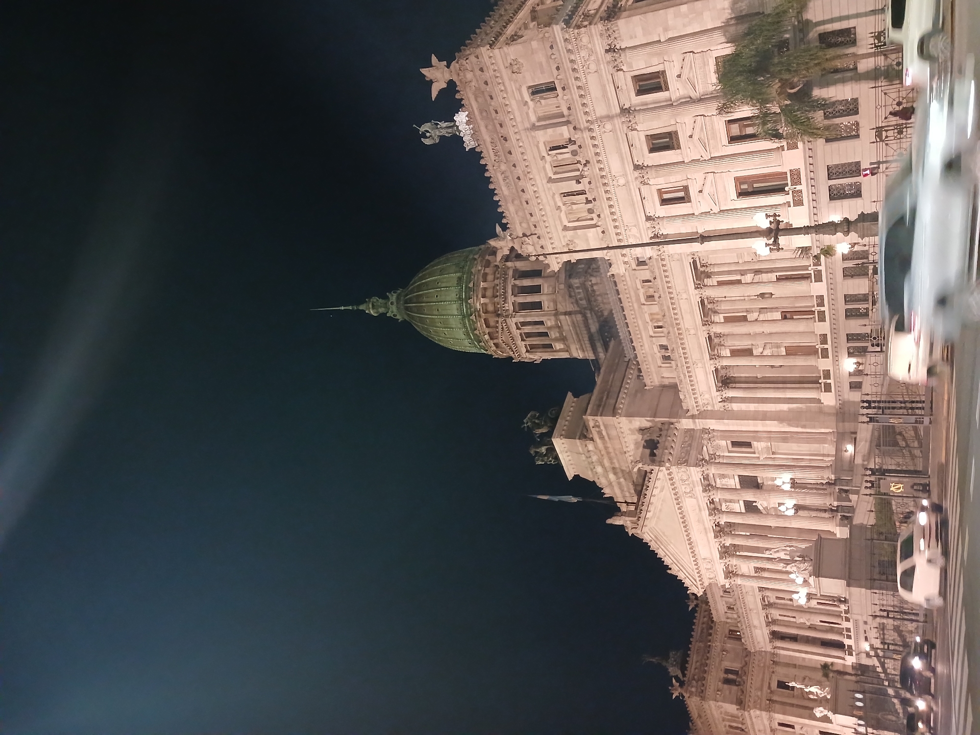 The image shows the Palace of the Argentine National Congress in Buenos Aires, Argentina at night. The building is illuminated and detailed with its architectural features, including columns, windows, sculptures, and a green dome. Streetlights and passing vehicles are visible in the foreground along the street. The sky is dark.