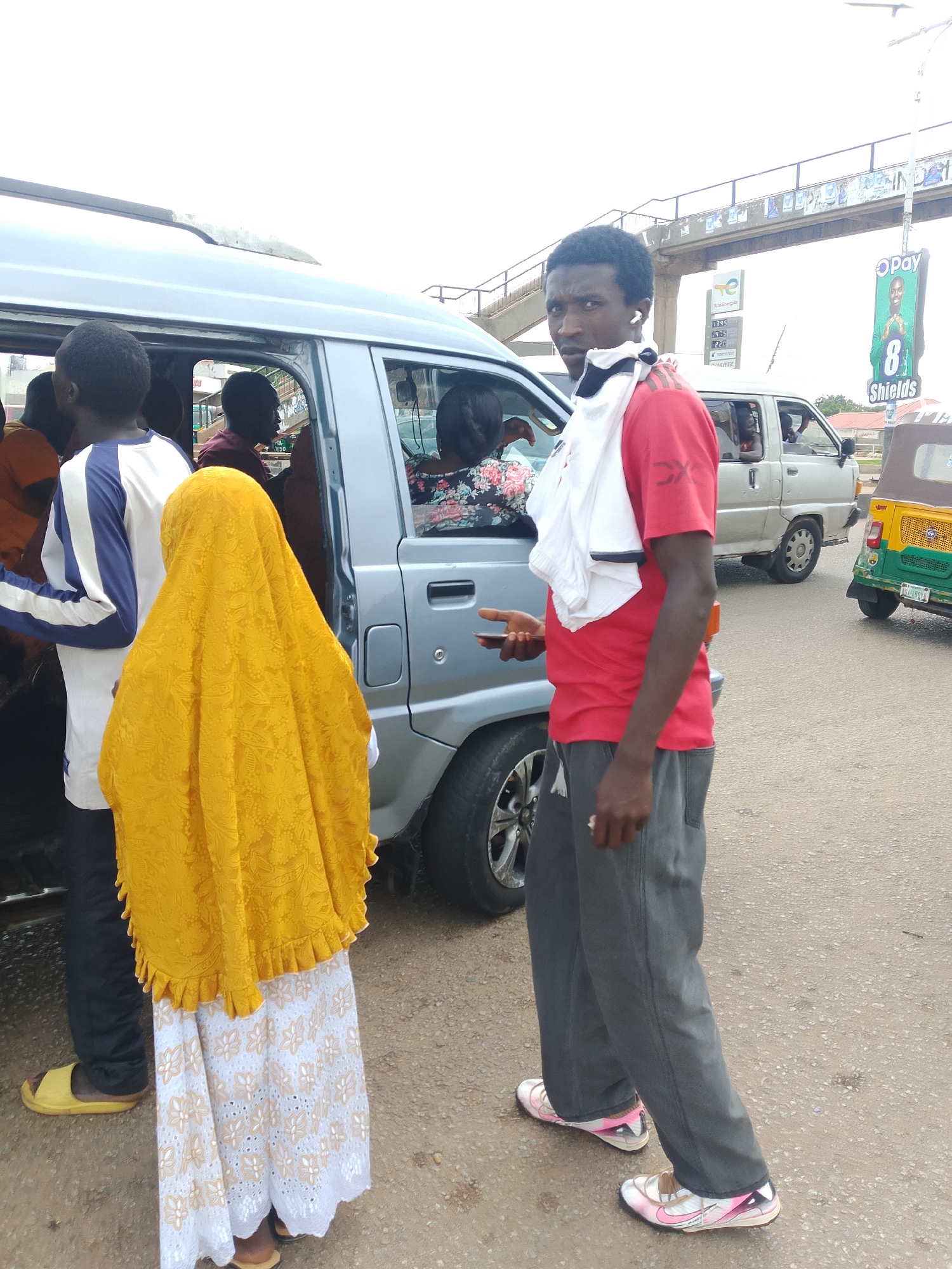 A man in a red shirt and grey pants is standing on a street next to a van. He is looking at the camera with a serious expression and has a white cloth draped over his shoulders. Another person wearing a yellow headscarf and a white and gold patterned skirt is standing to his left. Several other people are inside the van. In the background, there is a pedestrian overpass, a gas station sign, and a billboard with an advertisement. The sky is overcast.