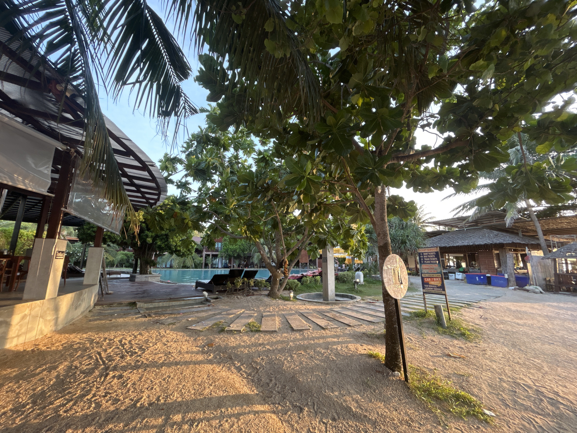 The image depicts a sun-drenched beachside area, likely a resort or a similar establishment, in the Ko Pha-ngan District of Thailand. The foreground is dominated by a sandy path illuminated by the warm, low-angled light of what appears to be late afternoon or early morning sun, casting long shadows from the surrounding vegetation and man-made structures.
To the left, a covered walkway or open-air building with a corrugated roof and support pillars is visible, featuring a sign that reads "Toilet". Further into the scene, a swimming pool with clear blue water is bordered by a wooden deck. Several dark-colored lounge chairs are arranged on the deck, some occupied by individuals who appear to be relaxing, with one person lying down.
Dominating the mid-ground is a large tree with lush green foliage, casting shade over the sandy area and the wooden pathway that leads towards the pool. A circular water feature or fountain is situated near the base of this tree. To the right of the tree, a signpost with a circular emblem and a multi-panel sign is positioned. The multi-panel sign displays text related to services, including "TICKET SERVICE," "BUS TICKETS," "TRAIN TICKETS," "AIRPORT TAXI," "SIGHTSEEING TRIP," and "THAI COOKING CLASS," among others.
In the background, additional resort buildings with thatched roofs and palm trees are visible, suggesting a tropical environment. The overall scene conveys a tranquil and relaxing atmosphere typical of a beach resort destination.