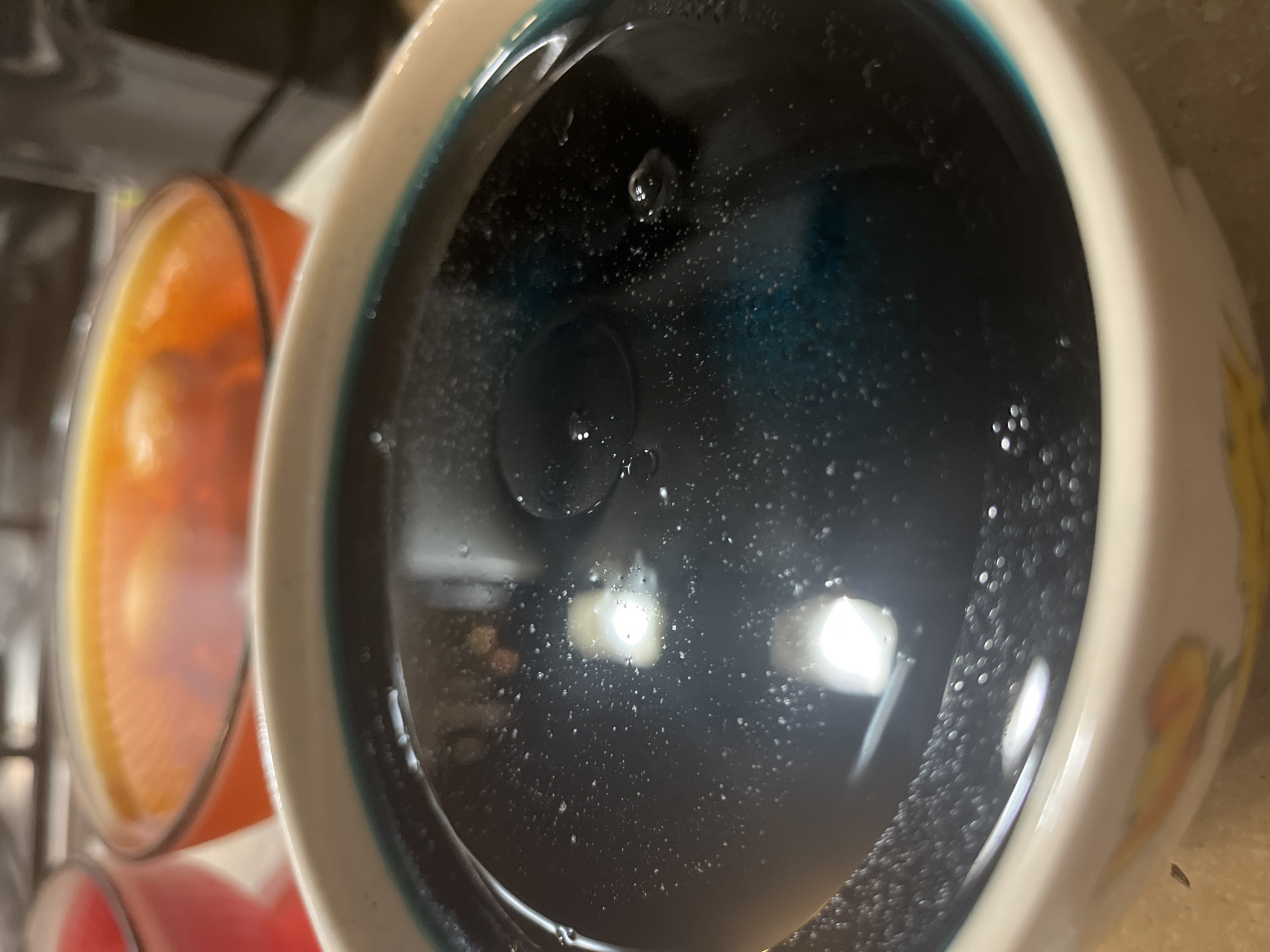 A close-up, canted perspective image depicts a white mug filled with a dark blue liquid, likely a dye solution. The mug's rim is lined with a thin blue band. Bubbles adhere to the inside surface of the mug. Reflections of light sources are visible in the liquid. Adjacent to the primary mug, parts of an orange and red container are visible. The background is blurry and out of focus. The image gives no indication of location in Chaikovsky, Russia.