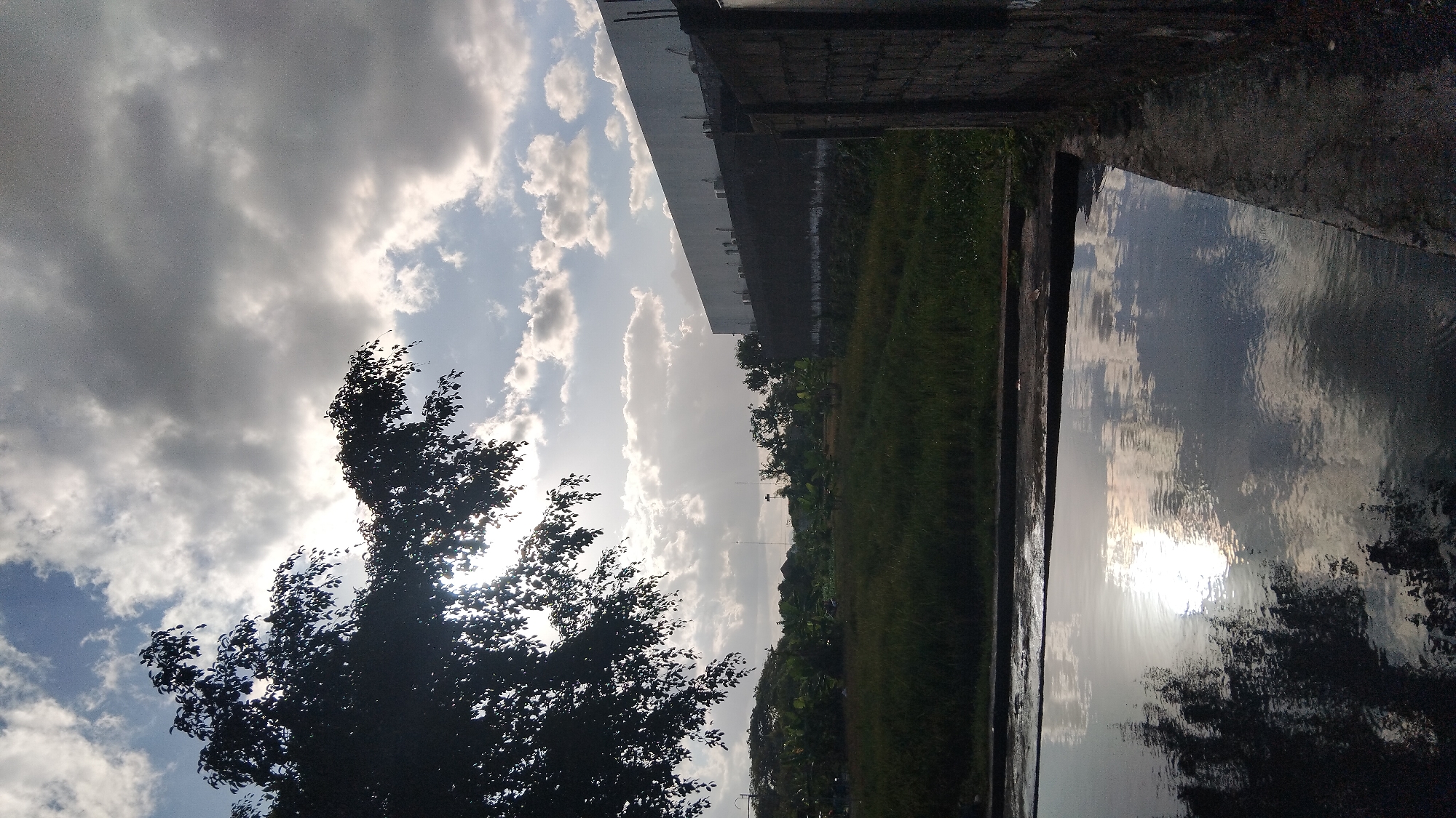 The image depicts an outdoor scene, likely in Ngaglik, Indonesia, characterized by a cloudy sky with visible sunbeams breaking through. In the foreground, a body of water reflects the sky and a silhouetted tree. To the left, a large tree with dark foliage partially obstructs the view. The middle ground features a grassy field with scattered vegetation. In the background, a long, light-colored building with visible air conditioning units stands behind a darker, lower structure. To the right, a brick wall is partially visible. The overall impression is one of a somewhat overcast day with a mix of natural and man-made elements. There are no people or animals visible in the image.