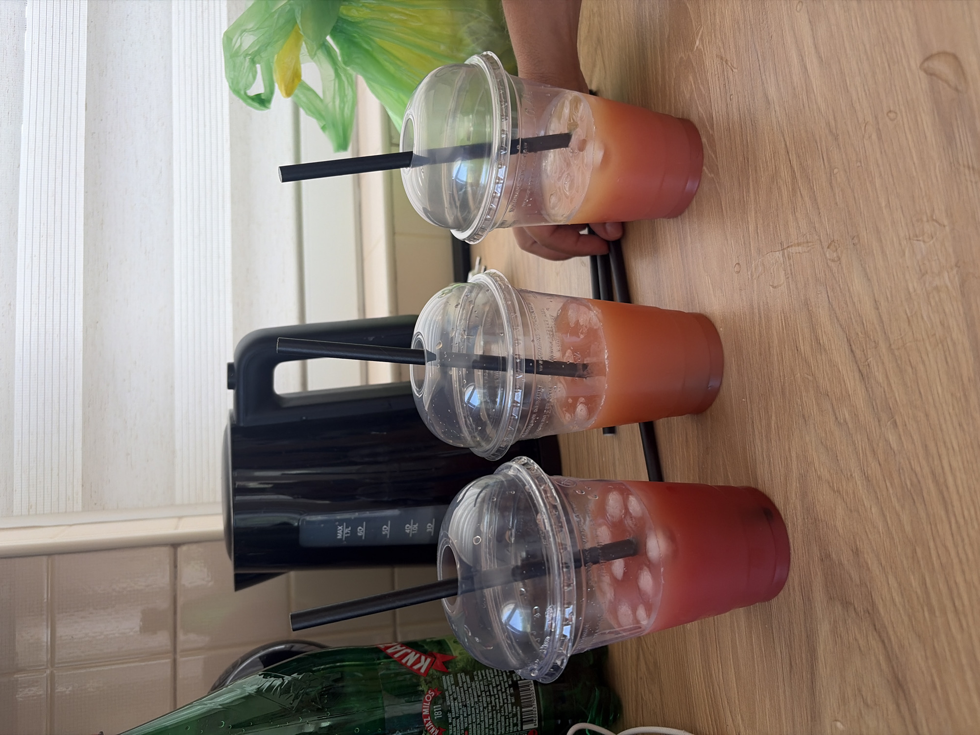 In a kitchen setting, on a wooden countertop, sit three disposable plastic cups filled with iced beverages. Two cups contain a reddish-pink drink, while the third holds a lighter pinkish-orange drink. Each cup has a clear, domed lid with a black straw inserted. To the left, a green glass bottle with a label partially visible, possibly containing water, stands upright. Behind the cups, a black electric kettle is situated. In the background, a light-colored tiled wall and window blinds are visible. To the right, a green plastic bag is crumpled, and a person's hand is partially visible reaching towards the cups. The overall scene appears to be a casual, domestic environment, possibly in the city of Lastva Grbaljska, Montenegro, given the context.