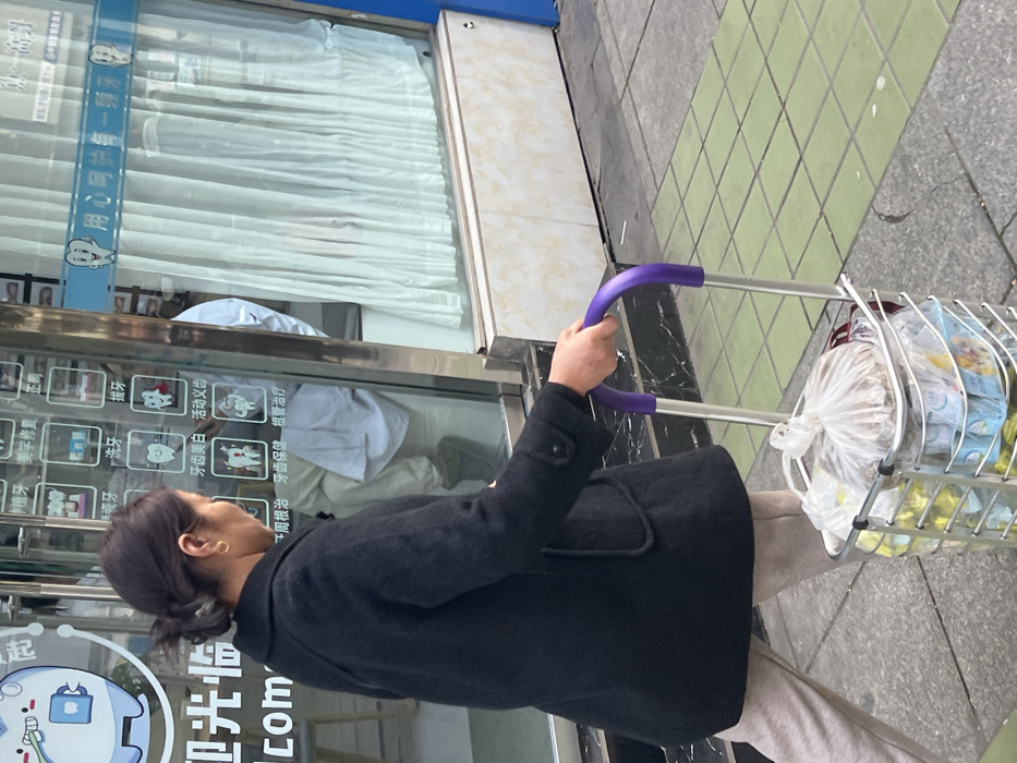 An outdoor, eye-level photograph shows a woman walking from left to right on a paved sidewalk in Jiangjin, China. The woman, viewed in profile, has dark hair tied back and wears a black coat over light-colored trousers, with a gold earring in her visible ear. She is pushing a silver-colored shopping trolley with purple handlebars, which contains multiple clear plastic bags filled with various packaged items, including what appears to be groceries. To her left is the glass storefront of a dental clinic, indicated by smiling tooth logos and Chinese characters on the window advertising services such as implants, whitening, and extractions. Through the glass, a light-colored curtain is visible on the right, and reflections on the left partially obscure what appears to be dental equipment or a chair. The sidewalk consists of light grey and green square tiles, with visible cracks in the grey sections. The lower facade of the building beneath the window is a light-colored, mottled panel.