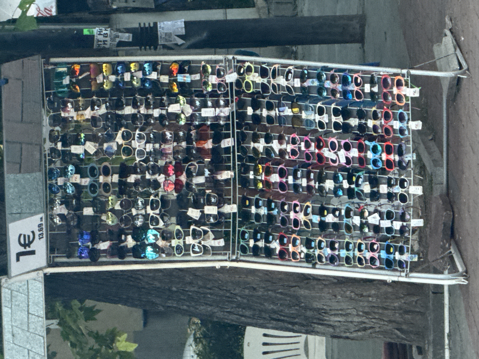 An outdoor rack of sunglasses is displayed for sale on a sidewalk next to a tree in the city of Unknown, Unknown. The rack is tall and narrow, made of white metal, and is filled with rows of sunglasses. The sunglasses are of various styles and colors, including black, white, blue, pink, red, green, and yellow lenses. Some sunglasses have reflective lenses. A sign at the top of the rack reads "1€" and "13.69 лв.". The sunglasses are attached to the rack by small tags. In the background, a tree trunk is visible, and further in the background, a white chair and a pole with some papers attached are visible. The ground in front of the rack is paved with brick and stone. The lighting suggests it is daytime.