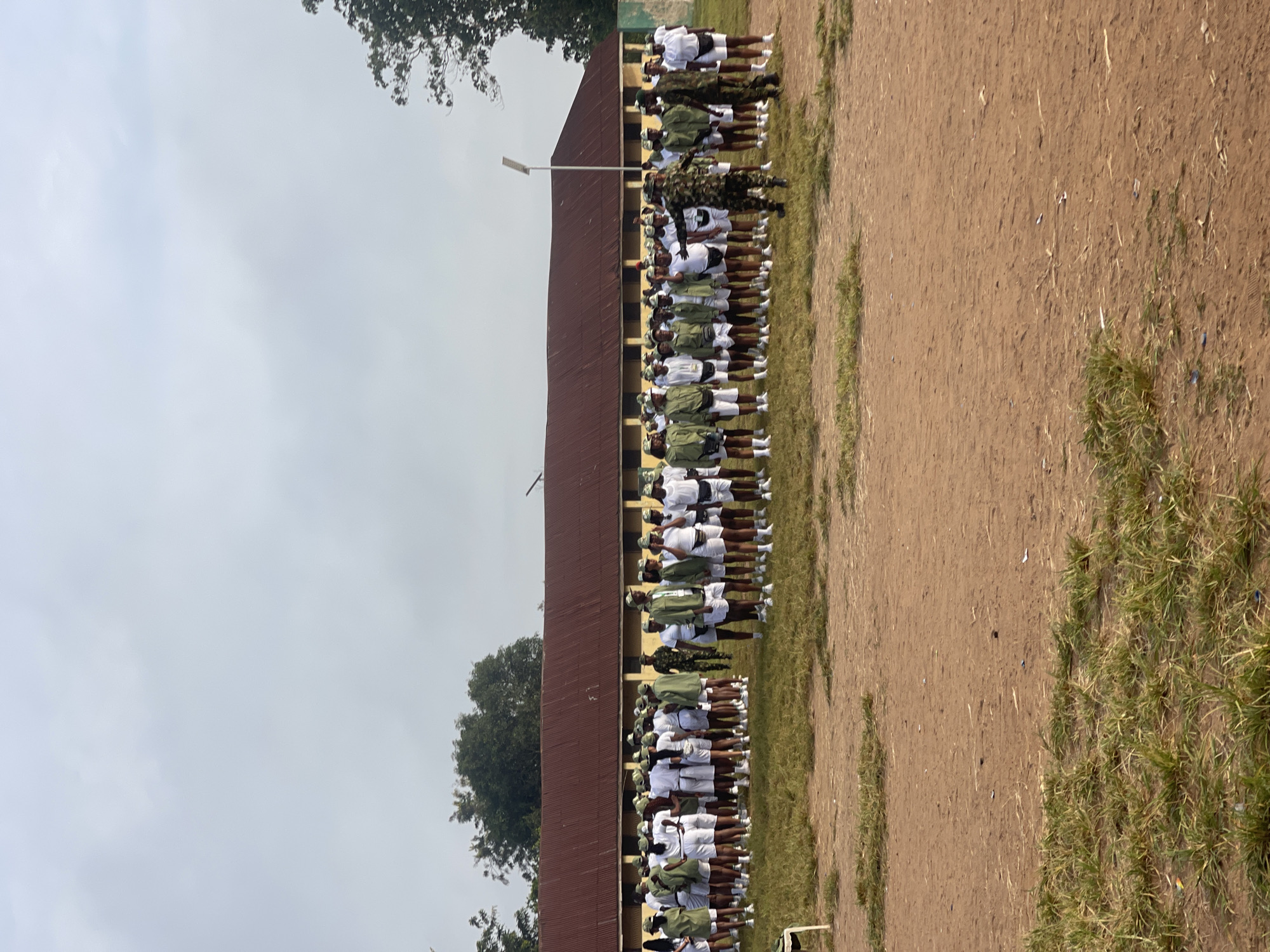 The image captures a group of individuals, likely uniformed cadets or participants, standing in formation on a grassy area in front of a long, yellow building with a corrugated iron roof. The majority of the group are dressed in white shorts and shirts with olive green vests and caps, suggesting a uniform. Several individuals in the foreground are wearing full olive green camouflage uniforms and appear to be instructors or supervisors, one of whom is in motion, possibly demonstrating a drill. The environment appears to be an outdoor training or assembly ground, with a dirt path visible in the foreground and trees in the background. The sky is overcast. Based on the context of similar images from this location, this scene is likely at the NYSC Orientation Camp in Ise - Ekiti, Nigeria, a common site for national youth service orientation programs.