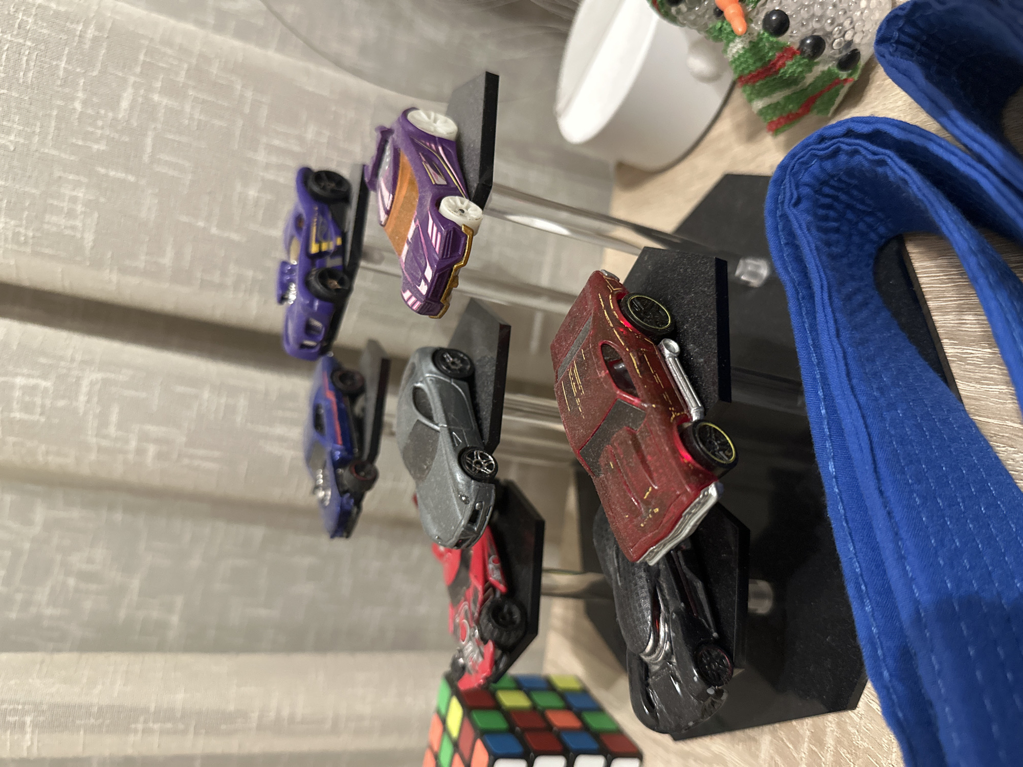 A collection of toy cars is displayed on multiple black stands, arranged at varying heights. The cars are predominantly sports car models in colors such as blue, purple, silver, red, and black. To the left of the cars, a Rubik's Cube is partially visible. In the background, a white textured curtain hangs. To the right of the cars, a white circular object and a small decorative snowman wearing a scarf are present. In the foreground, a blue fabric object, appearing to be a belt, is laid out. The overall scene is indoors, with artificial lighting.
