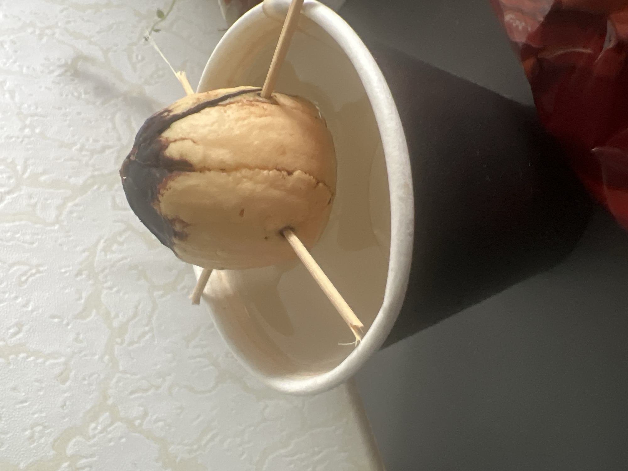 An avocado pit, suspended by three toothpicks, is submerged in water within a dark brown paper cup. The pit has a dark, possibly chocolate, residue on its upper portion. The cup sits on a dark, flat surface, with a textured, pale wall in the background. The scene is indoors and appears to be a home setting. The image does not contain any recognizable landmarks or elements that would indicate its specific location within the city of Pskov, Russia.