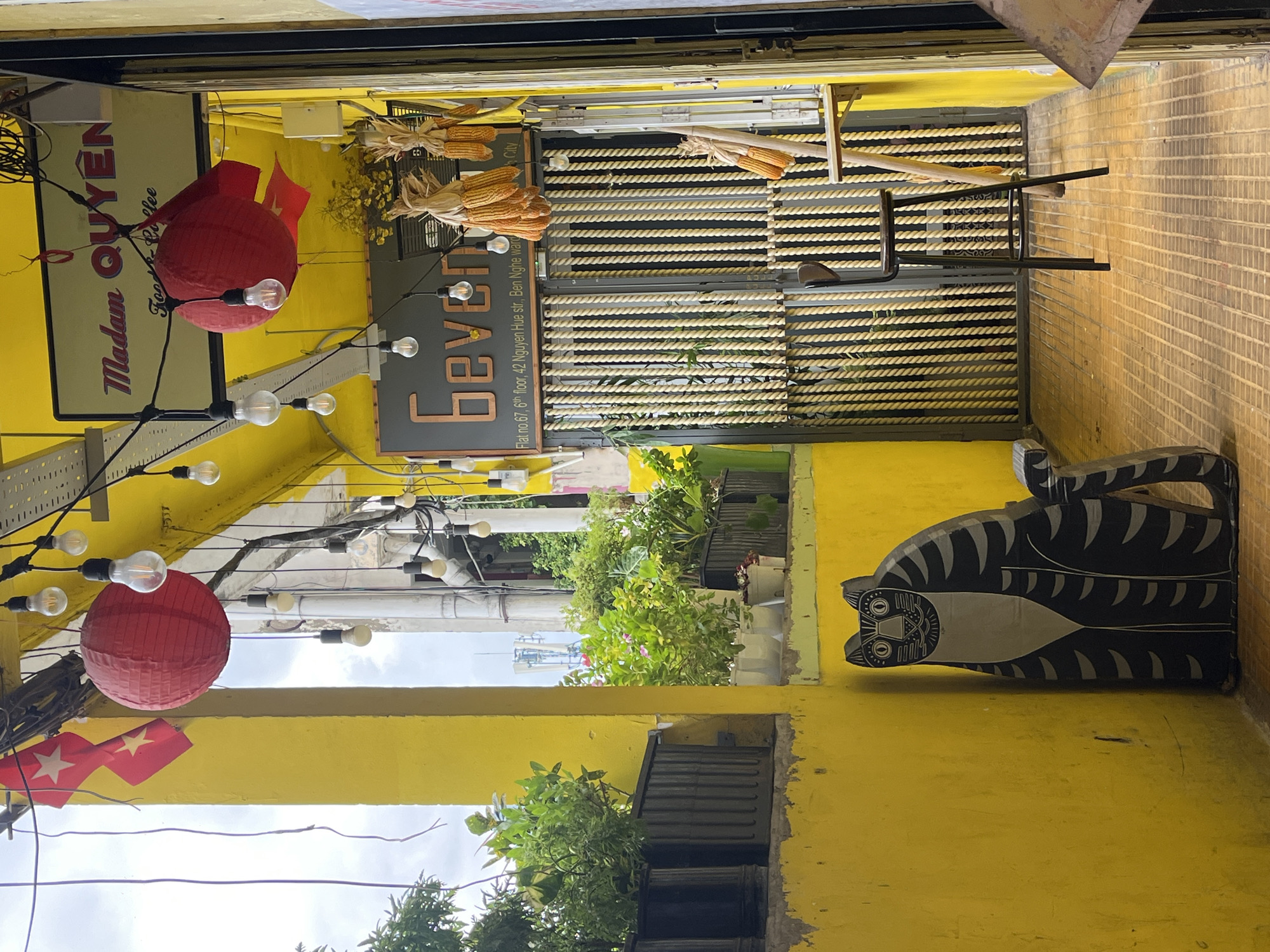 The image depicts the entrance of "Madam Quyên Food & Coffee," located at Flat no. 67, 6th floor, 42 Nguyen Hue street, Ben Nghe Ward, District 1, Ho Chi Minh City, Vietnam. The scene is characterized by a vibrant yellow facade and a covered walkway illuminated by a string of bare light bulbs. Two red paper lanterns hang prominently, one near a sign that reads "Madam Quyên Food & Coffee" and another slightly further down. Below this sign, another sign displays the name "Geven" in stylized lettering. Hanging from above are bunches of dried corn, adding a rustic element.
On the left side of the walkway, potted plants are arranged on a ledge against the yellow wall. In the foreground, a black and gray cutout of a cat is positioned near the wall. The walkway floor is paved with yellow tiles laid in a grid pattern. A tall bar stool with a wooden seat and back is placed in the middle of the walkway, facing towards a gate made of vertically arranged ropes. The overall atmosphere is eclectic and welcoming, blending modern elements with traditional decorative touches.