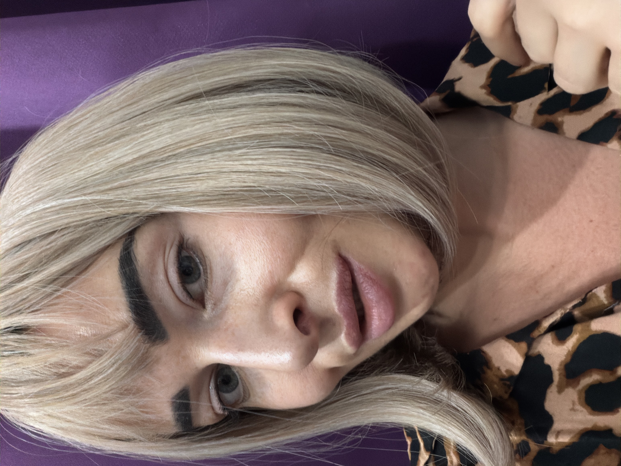 A close-up portrait features an individual with light skin, blonde shoulder-length bob hair, and prominent bangs. The person has dark, well-defined eyebrows, greenish-grey eyes, and light pink, slightly parted lips, forming a subtle smile. They are wearing a collared shirt with a brown and black leopard print pattern. The right hand, with light-colored fingernails, is visible gripping the collar of the shirt. The background is a plain, solid purple surface, suggesting an indoor setting. The image provides no visual cues to indicate a specific location within the city of Saint Petersburg, Russia.