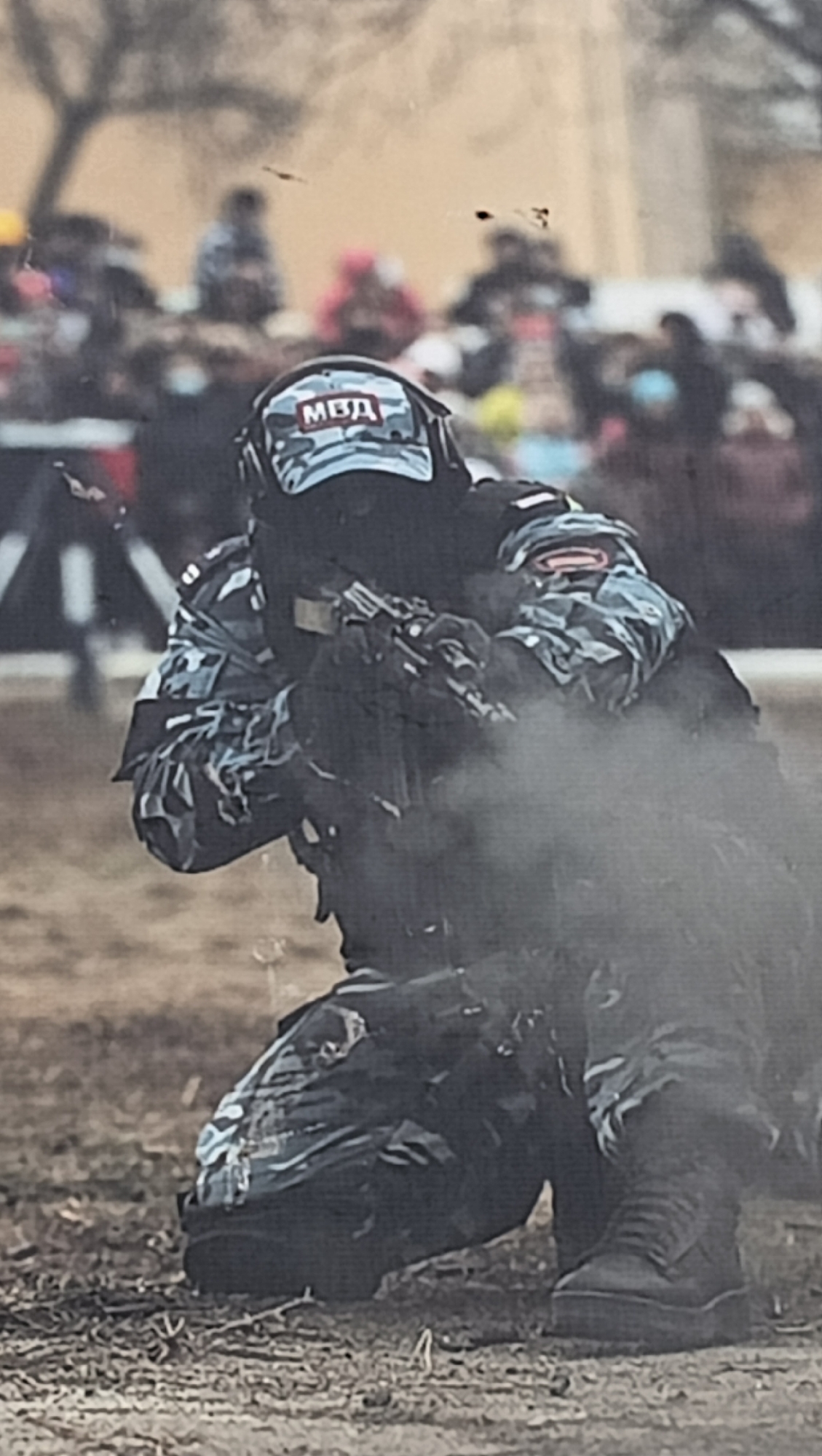 This daytime image, captured in Tiraspol, Moldova, depicts an individual in tactical gear kneeling and aiming a firearm. The subject is wearing a camouflage uniform in blue, grey, and white patterns, along with black tactical boots and gloves. Prominently displayed on their helmet or tactical headwear is the Cyrillic acronym "МВД" (MVD), which stands for Ministry of Internal Affairs.
The person is in a focused, ready stance, holding what appears to be a rifle or carbine. A cloud of smoke or dust billows around the front of the weapon and the individual, suggesting either a recent discharge or a special effect being used during an event.
The scene takes place outdoors on a dry, open field with sparse ground cover. In the blurry background, a crowd of spectators can be seen observing, alongside indistinct trees and foliage, indicating a public event or demonstration. The overall impression is that of a military or law enforcement exercise or display. The "МВД" designation is commonly used by internal affairs ministries in post-Soviet countries, including the Transnistrian region where Tiraspol is located. The weather appears clear.