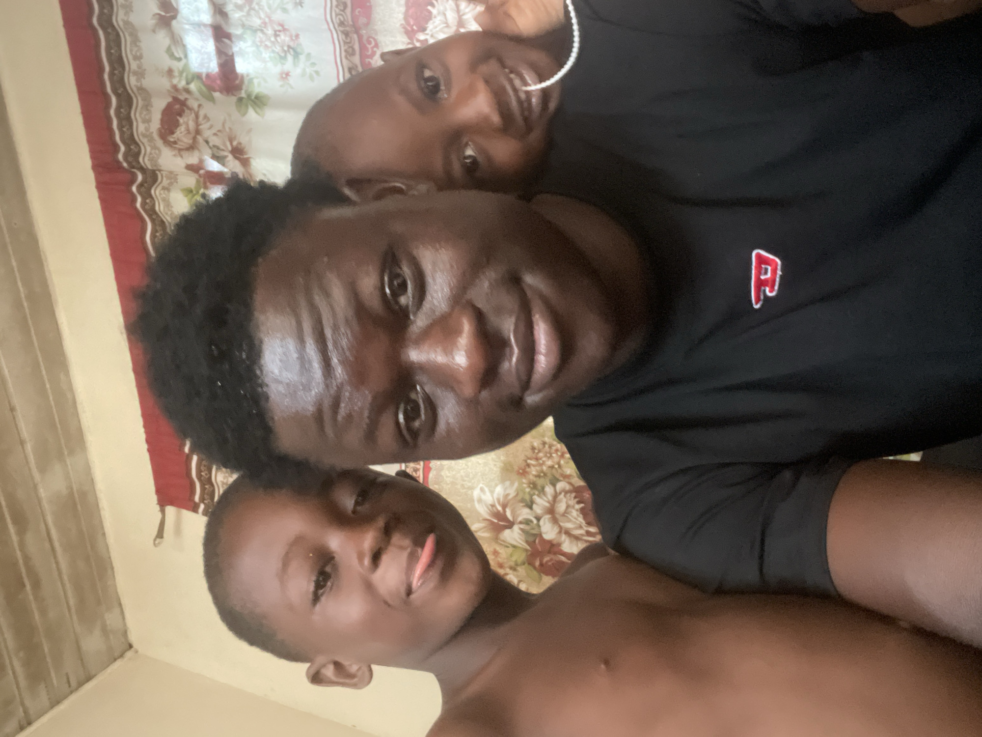 Three individuals are captured in a close-up, informal portrait. The central figure, likely an adult male, smiles directly at the camera. He is wearing a black t-shirt with a small, red "P" logo on the right chest area. Behind and above him is a second person, possibly another male, also smiling and wearing what appears to be a pearl necklace. In front of the central figure is a third individual, a younger male, shirtless, sticking his tongue out towards the camera. The background shows a floral patterned curtain in shades of red and beige. The overall lighting is natural. The location is presumed to be in Badagry, Nigeria, though no specific landmark is visible in the image.