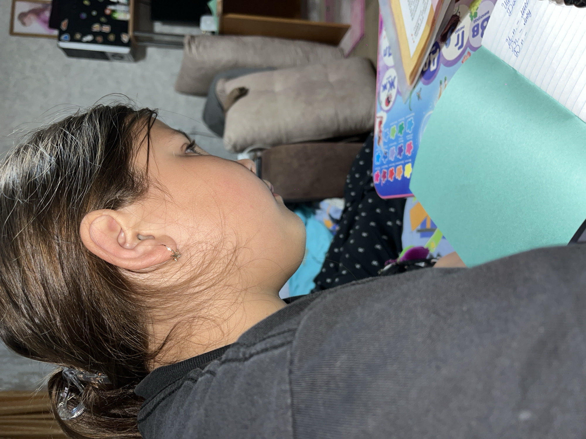 A girl with brown hair pulled back into a ponytail is seated at a table, possibly doing homework. She is wearing a dark grey t-shirt. She is looking to the right, towards a notebook and a green folder on a table. In the background, there is a patterned tablecloth and several pillows. A framed photograph is visible on a shelf in the background. The image's location in the city of Novopokrovskaya, Russia, cannot be verified.