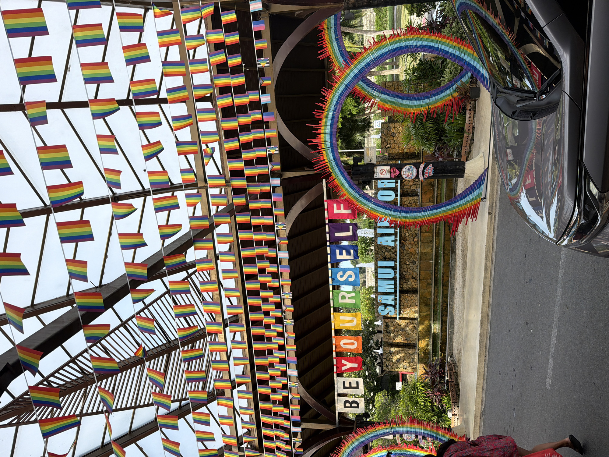 This media file is a photograph taken in **Ban Bang Rak, Thailand**. The scene is outdoors, possibly an airport or a public gathering space, decorated with numerous rainbow flags. The flags are small and strung together, creating a festive and colorful display. In the background, there are large rainbow-colored circular decorations and a sign with the text "BEYOURSELF SAMUI AIR". The image is dominated by the vibrant colors of the rainbow flags and decorations, suggesting a celebratory atmosphere, possibly related to Pride Month or a similar event promoting inclusivity. The time of day appears to be daytime, with natural light illuminating the scene. The weather seems clear and sunny. A black car is partially visible on the right side of the frame, reflecting the rainbow decorations. The overall impression is one of joy, celebration, and a welcoming environment.