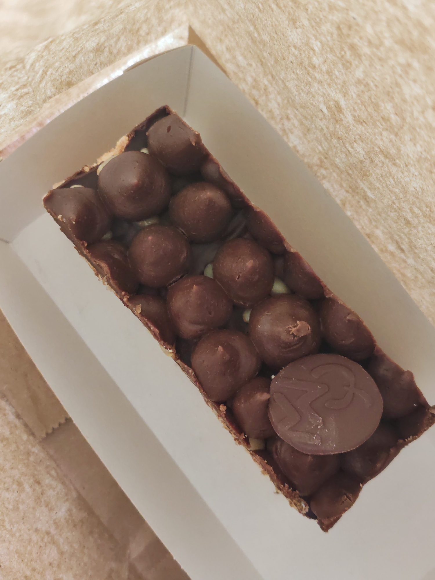 A rectangular chocolate dessert bar sits in a white cardboard box, which is placed on a textured, light brown surface, possibly paper packaging. The dessert is covered in numerous round, dark brown chocolate balls and has a thin, crispy base. A small, flat, round chocolate disk with a raised emblem, possibly a logo, is visible on top of the chocolate balls. The lighting suggests an indoor setting, likely a retail or food service environment. There is no indication of the specific location within Petropavlovsk-Kamchatsky, Russia.