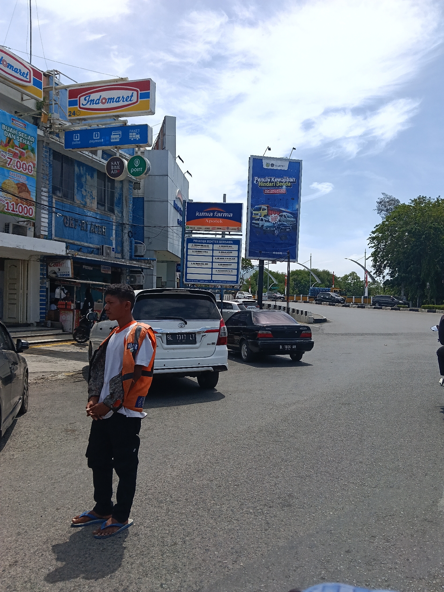 The image depicts a street scene in Banda Aceh, Indonesia, near an Indomaret convenience store and a Kimia Farma pharmacy. In the foreground, a man wearing a white t-shirt, an orange safety vest, and black pants stands on the asphalt road. He is looking to his left, with his hands clasped in front of him. Blue flip-flops are visible on his feet. To his right, a white Toyota Innova with a license plate reading "BL 1317 LT" is parked. A black sedan is parked behind the Innova, and its license plate reads "BL 1806 AW".
In the background, the Indomaret store has prominent signage, including "Indomaret" and a smaller sign with icons for "pulsa," "payment," "DEBIT," and "PAKET." A blue banner promoting "BUAH SEGAR DAN SEHAT" (Fresh and Healthy Fruit) with prices is also visible. Adjacent to Indomaret, the Kimia Farma pharmacy has its logo and contact information displayed. A large blue billboard advertising "Penulis Kewajiban Hindari Denda" (Writer of Obligations Avoid Fines) is situated further down the street. The sky is partly cloudy. The environment suggests a commercial area with moderate traffic.