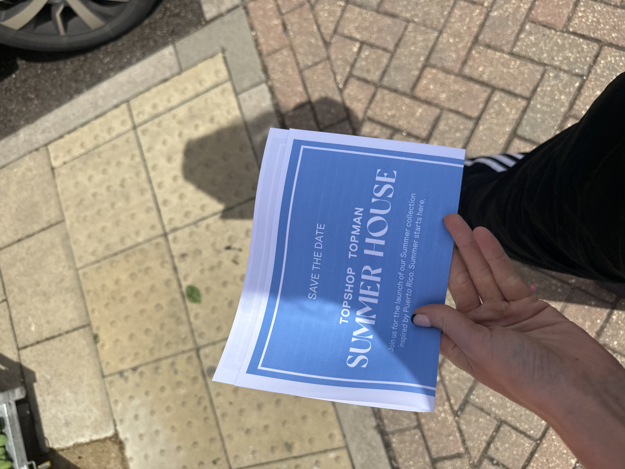The image captures a person holding a flyer or invitation in their hand. The flyer advertises a "Summer House" event by Topshop Topman, promoting the launch of their summer collection inspired by Puerto Rico. The person is wearing black pants with white stripes, and a hint of the sidewalk can be seen in the background, which appears to be made of paving stones. A car wheel is visible in the upper left corner of the image. The overall scene seems to be captured outdoors, potentially on a street or sidewalk in Tunbridge Wells, United Kingdom.