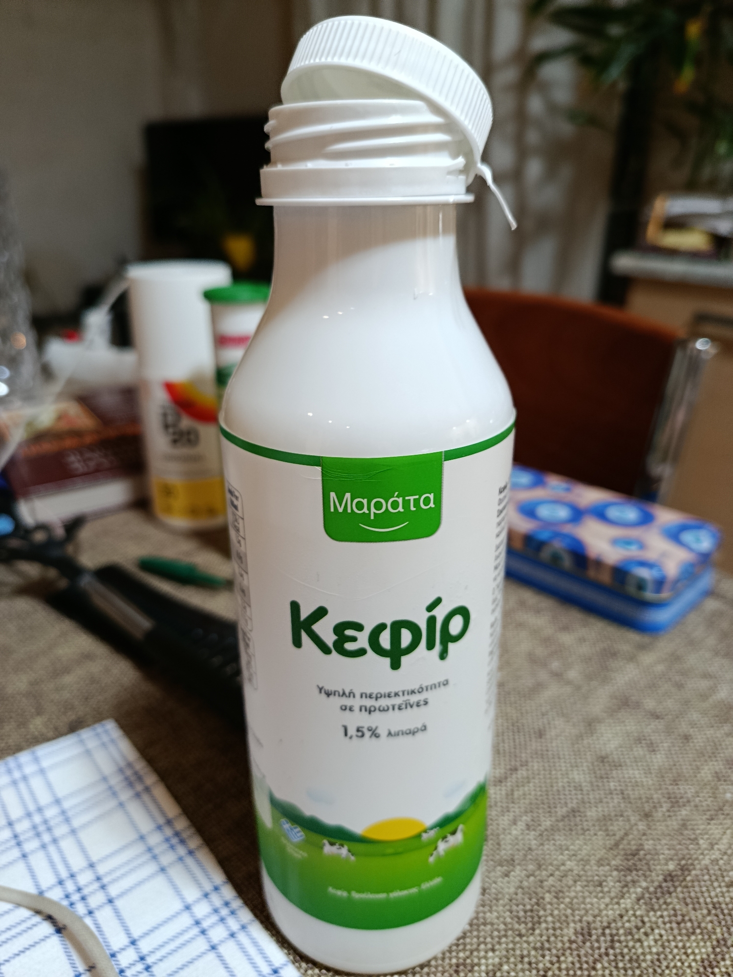 A white plastic bottle of kefir, with green and yellow branding, stands on a textured, beige surface. The label features green text that reads "Κεφίρ" and "Μαράτα", along with smaller Greek text and a depiction of a pastoral scene with cows and a sun. The bottle has a white screw-on cap with an attached flip-top lid. In the blurred background, other items are visible, including a white container, a book, and a chair. The scene appears to be indoors, possibly a kitchen or dining area. There is no discernible location in the city of Municipal Unit of Rhodes, Greece.