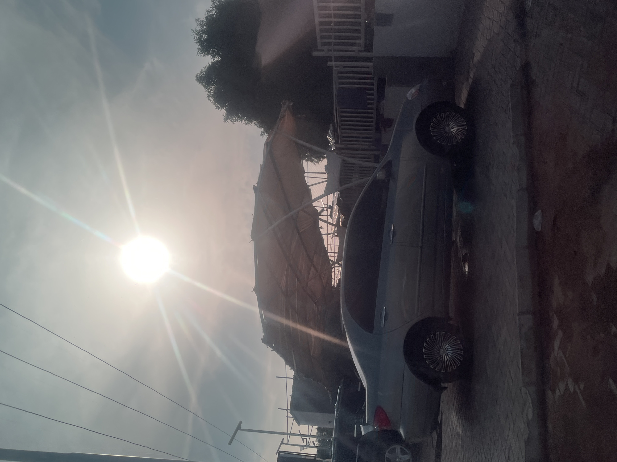 The image depicts a street scene, likely in Jalingo, Nigeria, under a bright, sunny sky. The sun is a prominent feature, casting strong rays and lens flares across the frame. Two cars are visible parked on a paved surface. One car, a light-colored sedan, is positioned in the foreground, angled towards the viewer. Its wheels and side profile are clearly visible. A second, darker vehicle is partially obscured behind the first. In the background, a makeshift structure with a thatched or canvas roof supported by poles is evident, suggesting ongoing construction or a temporary shelter. A tall tree with dark foliage stands to the left of this structure, its leaves silhouetted against the sky. The overall setting appears to be an outdoor urban or semi-urban environment, possibly a residential area or a commercial space. The time of day is clearly daytime due to the intense sunlight and shadows. There is no visible text in the image.