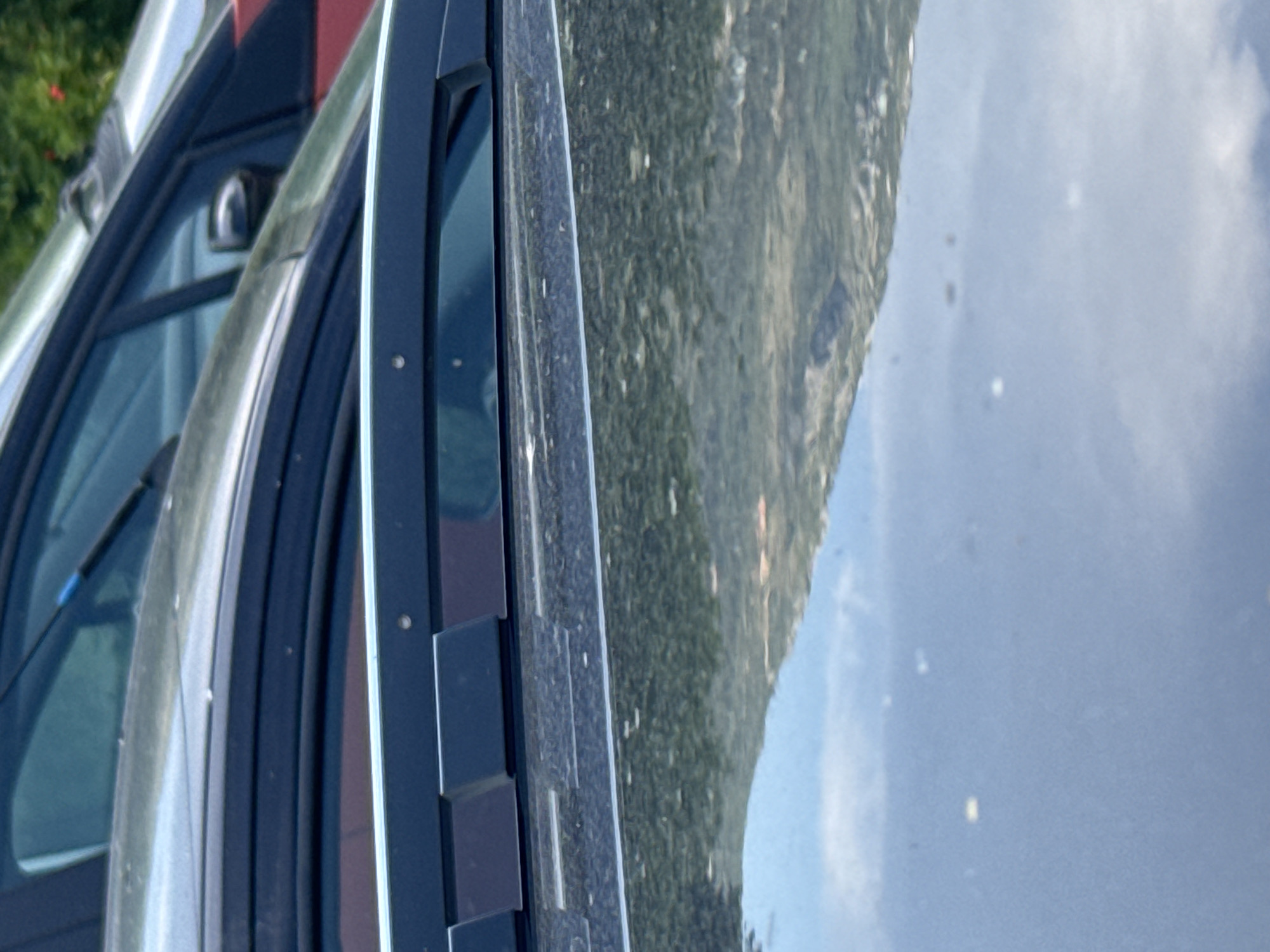 The image is a close-up, vertical shot of the side of a vehicle, possibly a car or van, parked outdoors. The vehicle's dark-colored frame and window are prominent in the foreground. A windshield wiper is visible along the top edge of the frame. Reflected in the vehicle's window is a landscape dominated by a steep, tree-covered mountain slope under a bright, partly cloudy sky. The scene suggests a daytime setting, likely during pleasant weather. The mountain's dense vegetation is a mix of green and lighter hues, indicating a natural, perhaps rural or mountainous area. The sky shows patches of blue and white clouds, with some bright spots that could be the sun reflecting or small, airborne particles. No people are visible. The image's perspective is from slightly above and to the side of the vehicle, focusing on the car's structure and the reflected scenery. The location context of Radanovići, Montenegro, aligns with the mountainous terrain depicted.