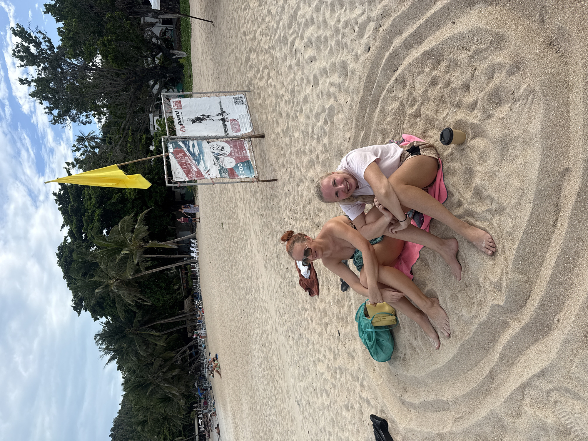 Two women are sitting on a sandy beach in the city of Ko Samui District, Thailand. The woman on the left is wearing sunglasses, a green bikini top, and has her hair pulled up in a bun. The woman on the right is wearing a pink t-shirt and shorts, with her blonde hair in a braid. They are both sitting on pink towels, with a green bag and a yellow purse next to the woman on the left. In the background, there are palm trees, other beachgoers, lounge chairs, and a yellow flag on a pole. There are also signs advertising parasailing and flyboarding. The sky is cloudy, with patches of blue sky visible.