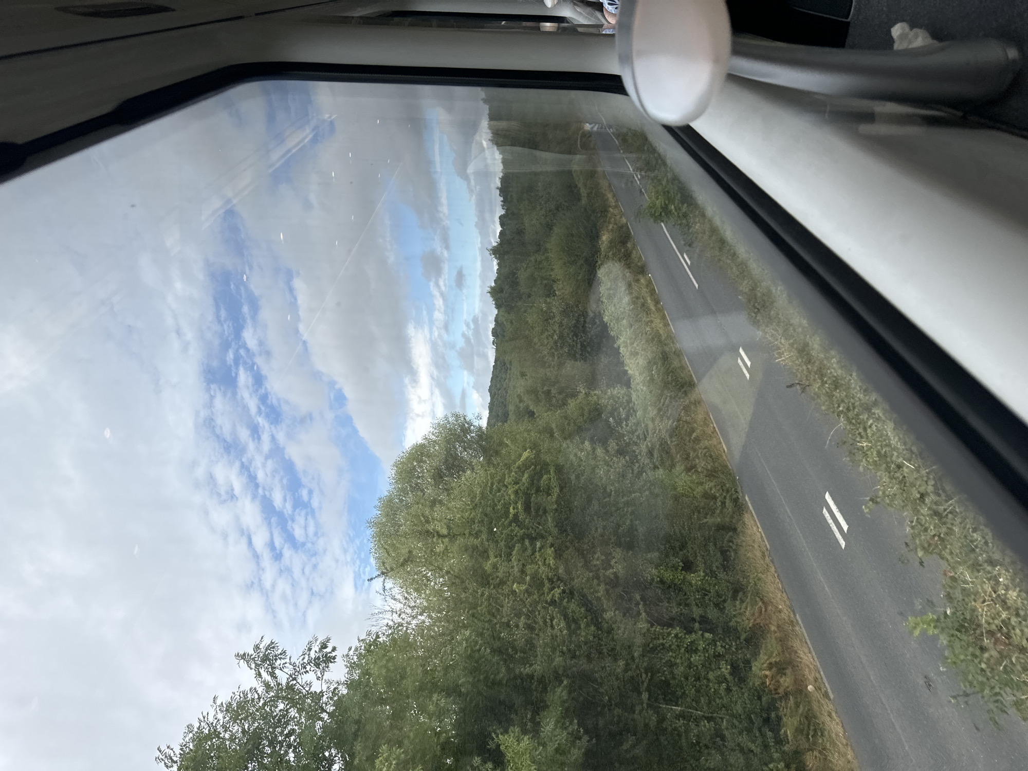 This image captures a view from a train window, looking out at a rural landscape. A two-lane road runs parallel to the train tracks, bordered by lush green trees and vegetation. In the distance, rolling hills are visible under a partially cloudy sky with streaks of blue. The interior of the train is partially visible in the foreground, with a white armrest and window frame. The scene appears to be in the town of Buckstown, Ireland, suggesting a journey through the Irish countryside.