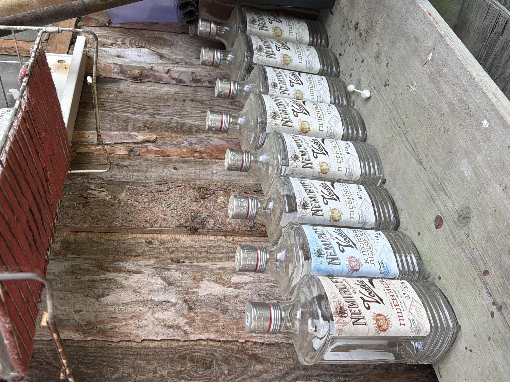 The image depicts a collection of ten empty glass bottles of Nemiroff vodka lying on a weathered wooden surface. The labels on the bottles indicate different varieties, including "Пшеница" (wheat), "Клюква ледяная" (cranberry ice), and "Original." Each label displays the vodka's volume as 0.7 liters and an alcohol content of 40%. The bottles are dusty and appear to have been discarded. The environment seems to be a rustic, possibly outdoor setting, indicated by the rough wooden planks and weathered appearance of the materials. There are no people visible in the image. Based on the vodka brand, which is produced in Ukraine, and the use of Cyrillic script on the labels, the location is likely within a country where this brand is distributed, but not necessarily Russia. The mention of Noviy, Russia, is not validated within the provided context.