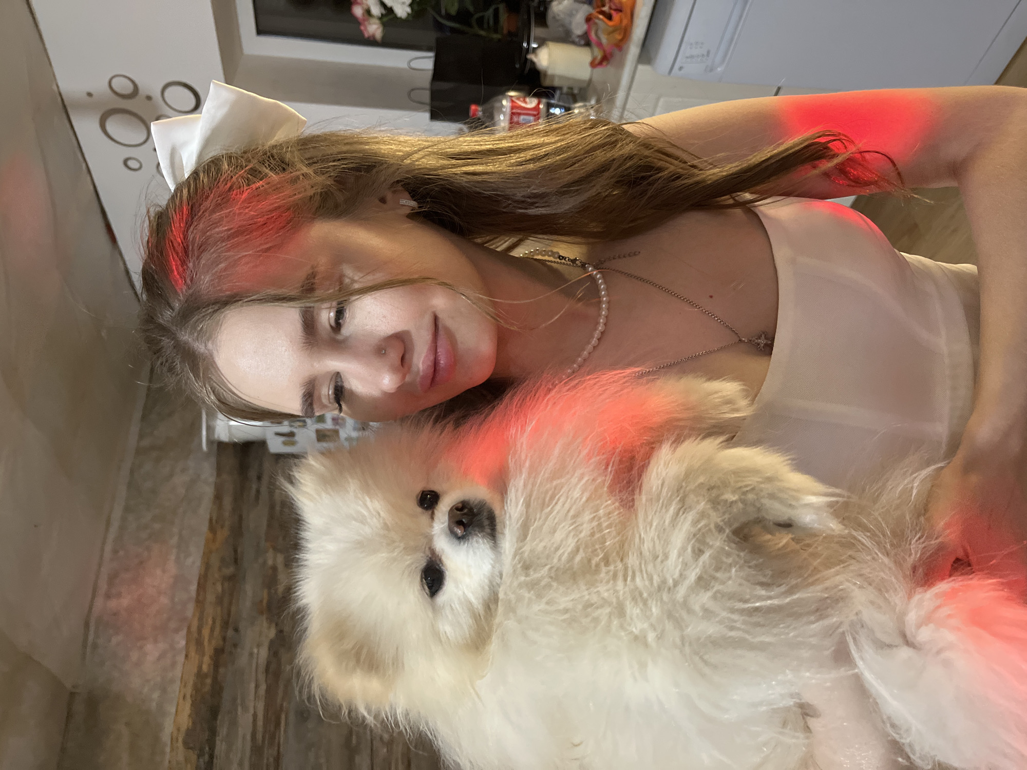 A woman with long, light brown hair and a large white bow in her hair is holding a small, fluffy white dog. The woman is wearing a light-colored dress and a pearl necklace with a cross pendant. Red light illuminates parts of her face, hair, arm, and the dog's fur. The background shows a wall with decorative circles, a wooden surface, and a glimpse of kitchen appliances and items. There is no indication in the image that this photo was taken in the city of Izhevsk, Russia.