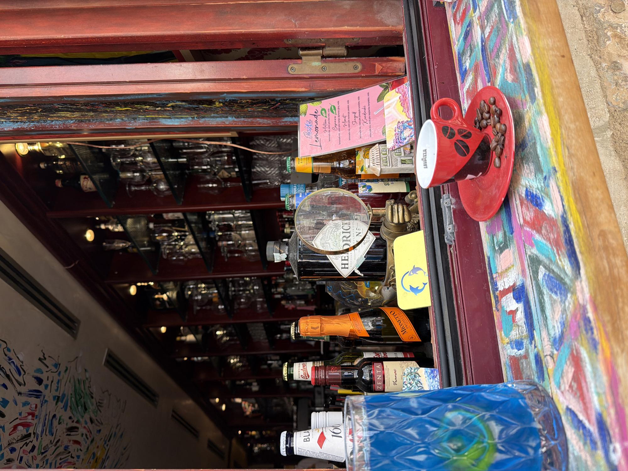 The image depicts a colorful, eclectic bar setup, likely located in the city of Budva, Montenegro, as evidenced by the context. In the foreground, a vibrant, multi-colored wooden counter displays a bright blue glass filled with a blue beverage on the left. To the right, a red Lavazza espresso cup sits on a matching red saucer, adorned with coffee beans. Behind the cup, a menu card with a pink background lists various lemonade options. The background reveals shelves stocked with numerous bottles of alcohol and glassware, suggesting a well-equipped bar. Bottles of various brands, including "BLAST," "Budweiser," "Zenta," and "MIONETTO PROSECCO," are visible on the counter and shelves. A bottle of Hendrick's Gin is also present. The overall impression is one of a lively and artistic establishment.