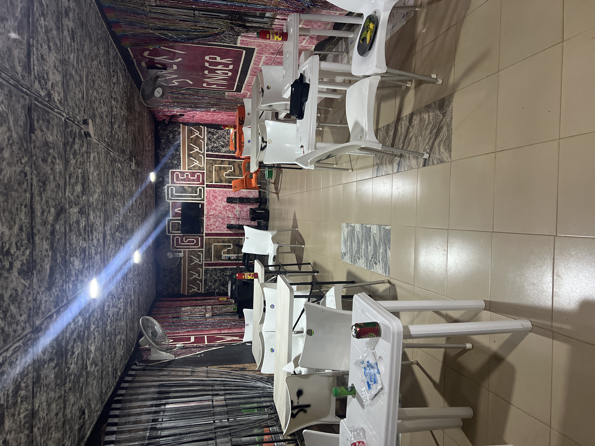 The image depicts the interior of an establishment, possibly a restaurant or bar, located in Nkpor, Nigeria. The space is furnished with numerous white plastic tables and chairs arranged in a dining or seating configuration. The walls are decorated with vibrant, colorful bead curtains and stylized lettering that reads "SWEET FINGER" on the right side and "GRACE" on the left. The ceiling is textured and appears to have exposed wiring and multiple light fixtures that cast a bright, cool-toned illumination. Two oscillating fans are mounted on the walls.
On the tables, several items are visible, including what appear to be plastic wrappers, a can of drink, and a bottle of sauce. The floor is tiled with square, light brown tiles, with some sections featuring a contrasting patterned tile inlay. The overall ambiance suggests a casual, possibly budget-friendly, establishment. There are no people visible in the scene.
