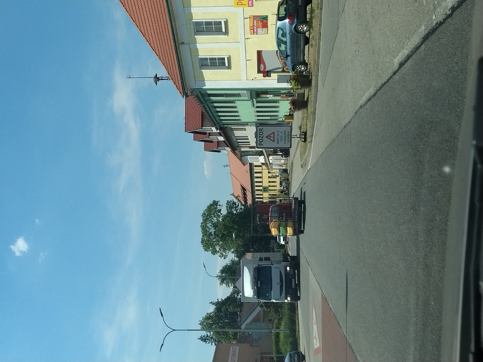 The image shows a street scene in Střelské Hoštice, Czechia. On the right side of the road, there is a row of buildings with red tile roofs and light-colored facades, painted in yellow and green. One building has a sign that says "ZDE V PRODEJI PLYNOVÉ LAHVE". A blue car is parked in front of this building. On the left side of the road, a white truck is driving towards the camera. Behind it, a red truck with a trailer carrying yellow and green objects is also moving away from the camera. A "POZOR" (Attention) sign is visible on the left side of the road, warning of a closure on a road marked with EV.Č. 17210 - 3. The sky is blue with a few white clouds. The road is asphalt.