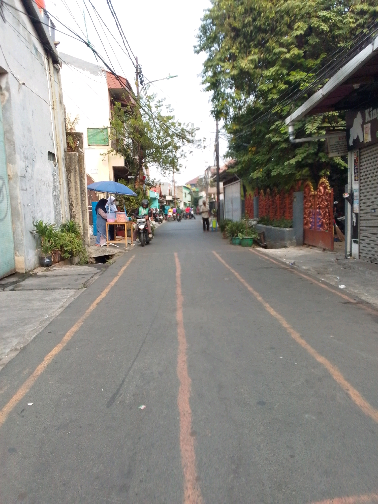 The image depicts a street scene in the city of East Jakarta, Indonesia. The perspective is from a low angle, looking down a paved road with orange lane markings. On the left side of the road, there is a building with a light-colored facade, and further down, a blue umbrella shades a small food stall with a wooden table. Two individuals, one wearing a headscarf, are standing at the stall, possibly serving or preparing food. In front of the stall, potted plants are visible. On the right side of the road, there are buildings with dark-colored awnings, and a decorative orange fence. Trees with green foliage line the sidewalk. Several motorcycles are present on the road, with one rider in a green helmet visible. A person is walking on the sidewalk in the background. The sky is overcast.