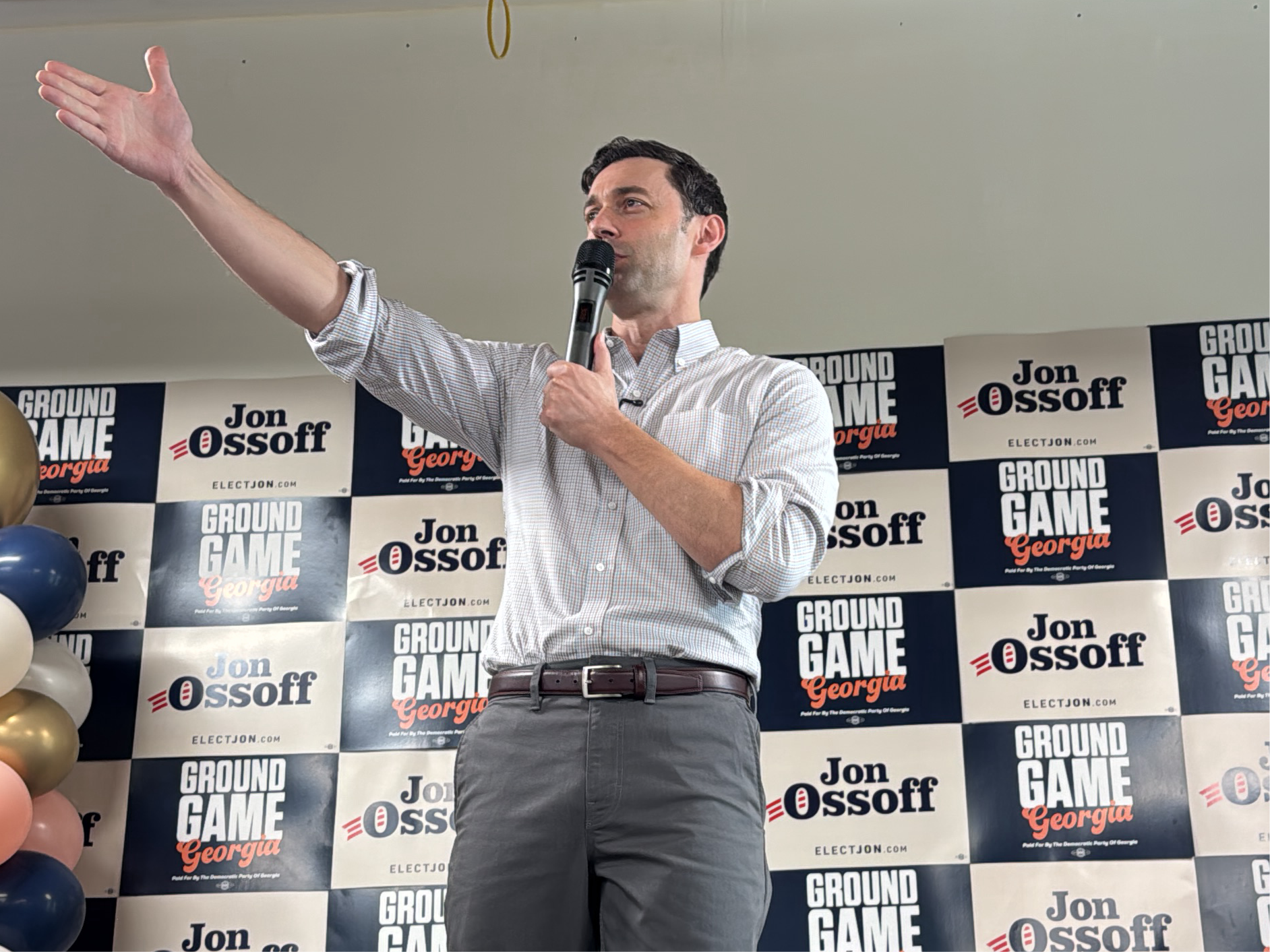 Here is a detailed description of the image:
**Scene:** The image appears to be taken at a political event or rally. The background is a repeating pattern of banners that read "GROUND GAME Georgia" and "Jon Ossoff" with the website "ELECTJON.COM".
**People:** The main subject is a man, presumably Jon Ossoff, who is standing and speaking into a microphone. He is dressed in a light-colored checkered shirt with rolled-up sleeves, gray pants, and a brown belt. He has dark hair and is gesturing with his left hand raised.
**Objects:**
* Microphone
* Banners with political branding.
* Balloons in the bottom left corner
**Actions/Interactions:** The man is speaking into a microphone, suggesting he is giving a speech or addressing a crowd. His raised hand indicates he is making a point or emphasizing something.
**Environment:** The environment seems to be indoors, possibly a hall or event space. The lighting is bright.
**Location:** While the explicit location in Decatur, United States, isn't visually identifiable in the image, the banners with "Georgia" clearly indicate the state, and it's reasonable to assume a connection to Decatur if that information is given.