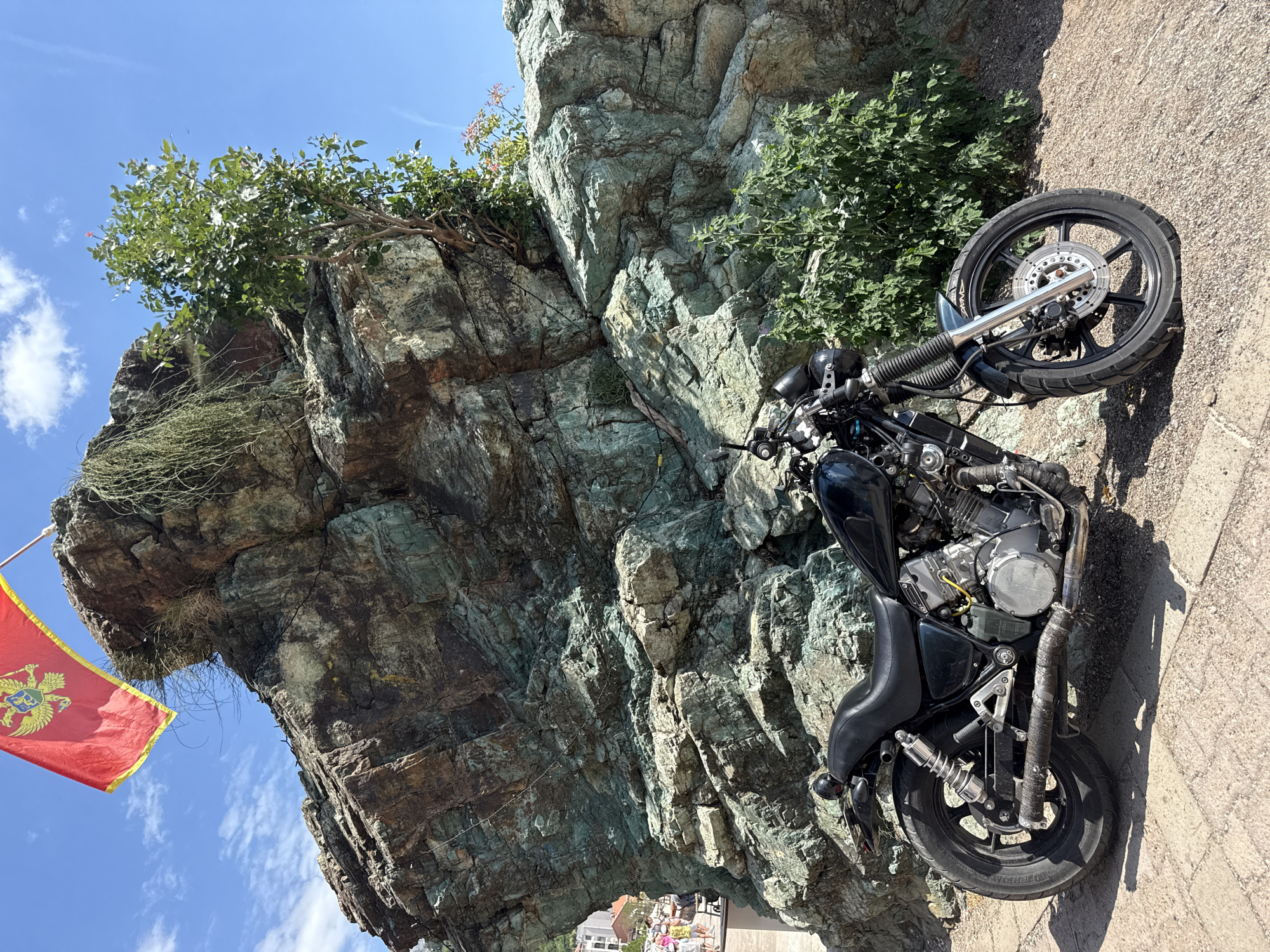 A black motorcycle is parked on a paved walkway in front of a large, jagged rock formation. A red flag with a golden emblem is flying from a pole atop the rock. The rock face has layers of green and brown minerals. Green foliage grows from crevices in the rock and a small tree with pink flowers is visible on the right side of the rock. The sky is blue with scattered white clouds. In the background, to the left, there are buildings and people walking. The scene appears to be in a coastal area, likely in Rafailovići, Montenegro, given the presence of the Montenegrin flag.