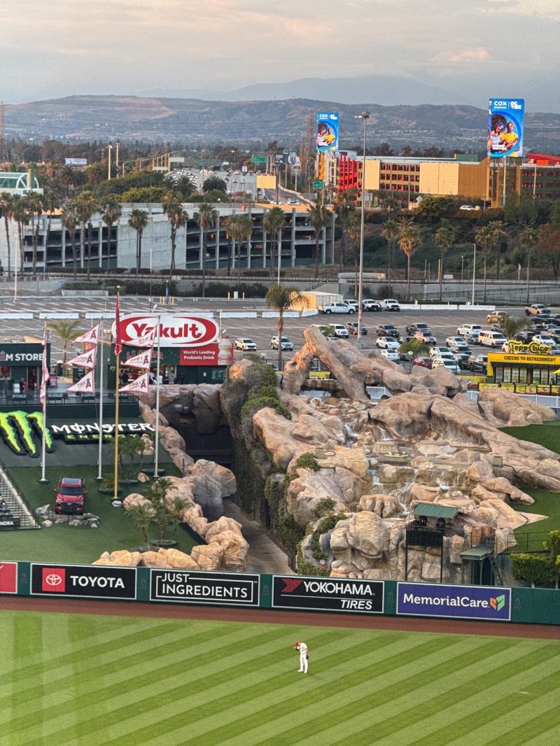 The image shows a baseball field in Anaheim, United States, likely taken from an elevated position within the stadium. The field is green with clear lines, with advertising banners visible along the edge of the field. The signage includes brands like Toyota, Yokohama Tires, Just Ingredients, and MemorialCare. There's a distinctive rock formation with a waterfall feature, partially obscured by greenery. A baseball player stands alone on the field. In the background, there's a parking lot filled with cars and buildings, with billboards displaying advertisements. The city skyline and distant mountains are visible in the background under a bright sky.