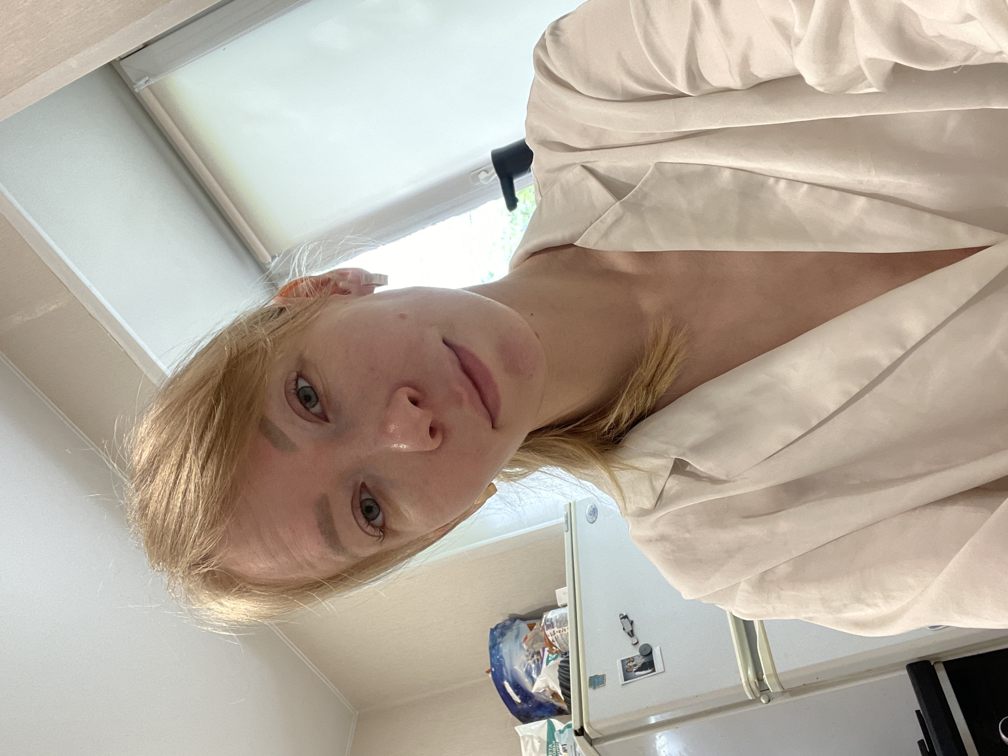 A selfie-style, slightly angled shot from above features a fair-skinned, blonde woman indoors. She has light eyes and a neutral expression. She's wearing a white blouse. Behind her, a white refrigerator is visible, adorned with magnets and a photograph. A package sits on top of the fridge. The setting appears to be a kitchen with light-colored walls and a window at the top of the frame. It is not possible to pinpoint the location as Perm, Russia from the visual information provided.