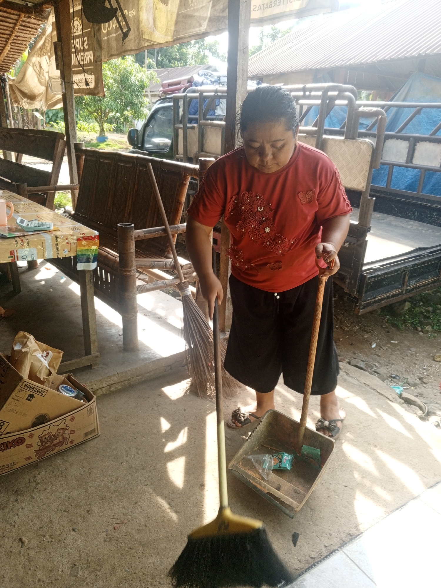 In Tanah Gara Hulu, Indonesia, a woman wearing a red shirt with intricate beadwork and black shorts is sweeping the concrete ground with a broom and dustpan. The scene appears to be an outdoor market or roadside stall, with bamboo furniture and a truck visible in the background. Sunlight streams down, casting shadows on the ground. A cardboard box filled with items sits to the left of the woman, and a table covered with various objects, including a calculator and a mug, is also present. The environment is rustic, with dirt and debris on the ground, suggesting a work-in-progress or a recently used space.