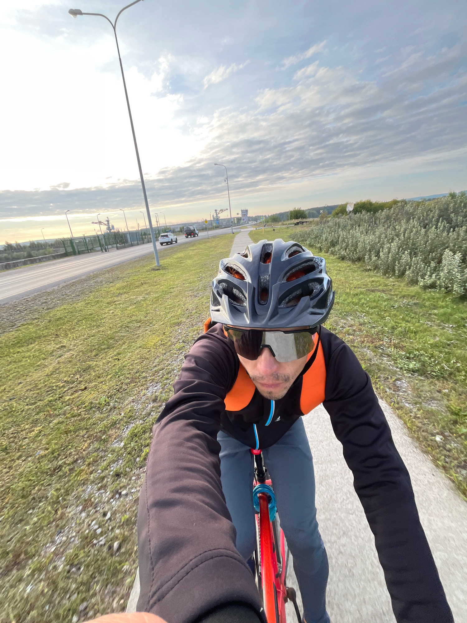 A man wearing a black cycling helmet, silver-tinted cycling glasses, a black long-sleeved jersey with an orange vest, and gray cycling pants is riding a red bicycle on a paved path. The path runs alongside a grassy area and a road with some traffic. In the background, there are industrial structures, trees, and a cloudy sky. The image appears to be taken in Chaikovsky, Russia, given the presence of industrial elements and the general landscape which is consistent with the region. The lighting suggests it is either early morning or late afternoon.