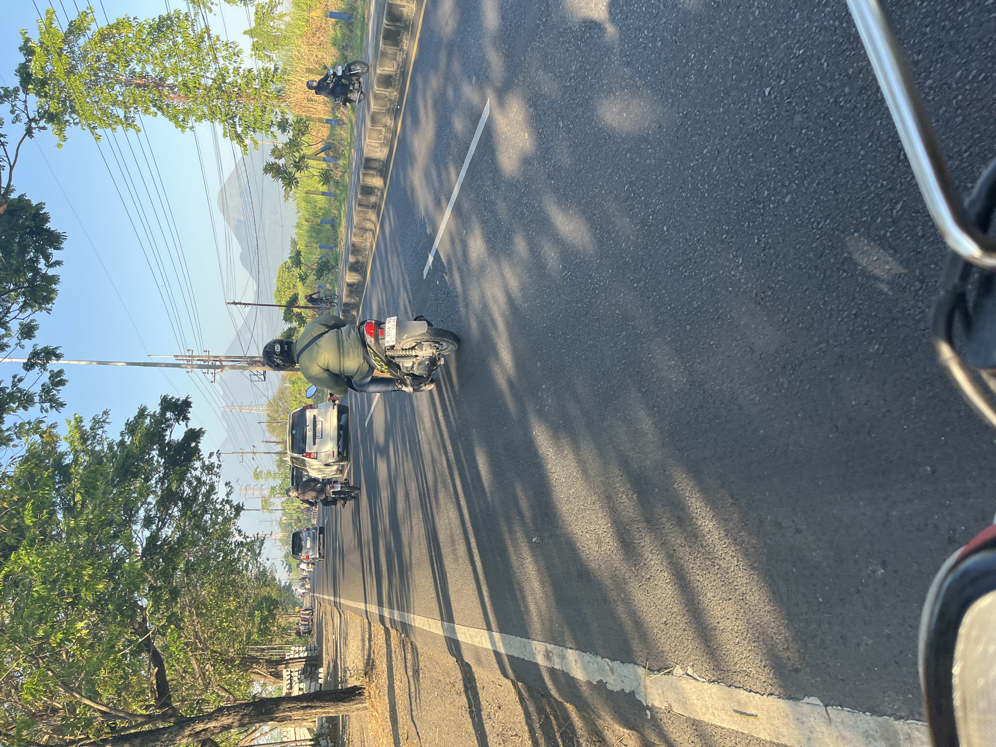 The image depicts a scene on a road in Siring, Indonesia, likely during the daytime given the bright sunlight casting long shadows. The perspective is from a vehicle moving along the road.
In the foreground, the handlebars of what appears to be a motorcycle or scooter are visible, suggesting the image was taken from the viewpoint of a rider. The road surface is asphalt with a white line marking the lane.
Several vehicles are present on the road. A person on a motorcycle, wearing a green jacket and a helmet, is in the right lane, moving away from the camera. Further ahead, a white car is visible. On the left side of the road, there are trees with lush green foliage, and a sidewalk or shoulder with some stacked materials. Power lines are strung across the sky.
In the middle ground, more vehicles are traveling in the same direction. A person on a bicycle is also visible. The background shows a hazy, mountainous landscape under a clear blue sky. The overall impression is a typical scene of traffic on a road in a semi-urban or rural area with natural elements.