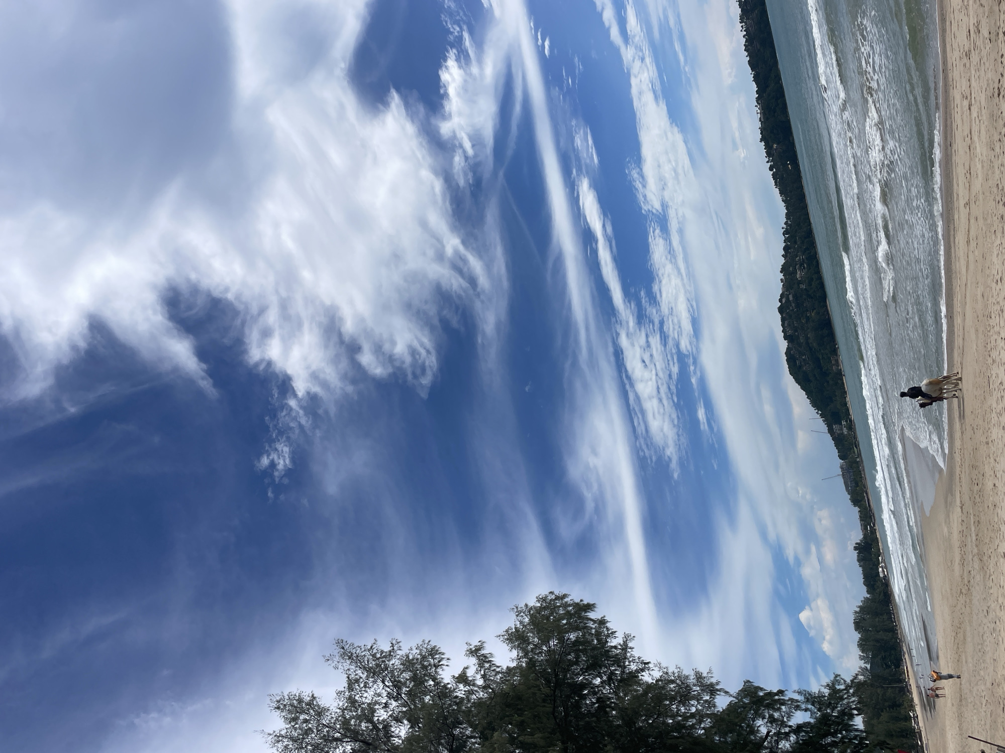 The image depicts a serene beach scene in Choeng Thale, Thailand. A vast expanse of blue sky with wispy white clouds dominates the upper half of the frame, creating a dramatic backdrop. Below, the turquoise ocean meets a sandy beach, with gentle waves rolling ashore.
In the foreground, a person is riding a light-colored horse along the shoreline. To the left, a lush green tree with needle-like leaves provides a natural frame for the scene. In the distance, a densely forested hill with scattered buildings rises from the coastline. Several other people are visible walking on the beach further down, some appearing to be tourists. The overall atmosphere is one of tranquility and natural beauty.