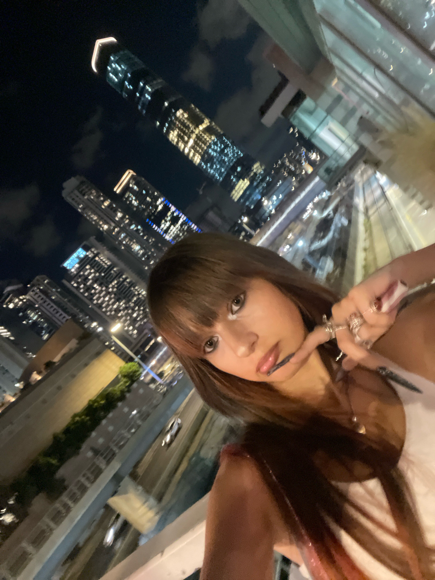A young woman with long brown hair and bangs is taking a selfie at night. She is wearing white clothing and has makeup on her face. She is wearing rings on her fingers and has long fingernails. In the background, there are several illuminated skyscrapers and city lights, suggesting an urban environment. The overall scene is a nighttime cityscape, possibly from a high vantage point like a balcony or rooftop. There is no visual information in the image to identify the location as Tel-Aviv, Israel.