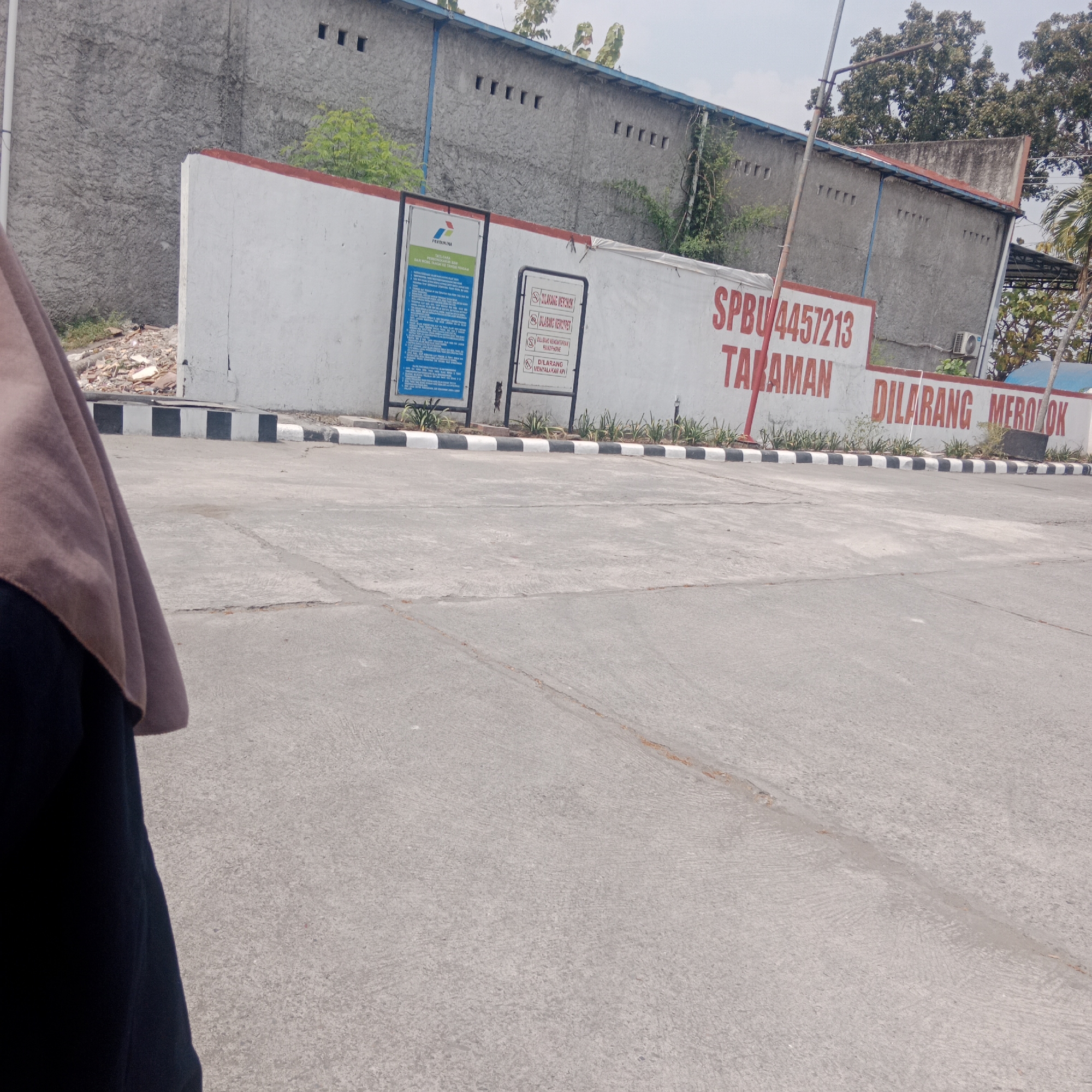 The image shows a concrete road in front of a white wall with red lettering. The wall has several signs attached to it. To the left, there is a large blue sign with text in Indonesian and a Pertamina logo. Next to it is a smaller white sign with red text and symbols indicating prohibitions. To the right, large red text on the white wall reads "SPBU 4457213 TARAMAN DILARANG MEROKOK" which translates to "Gas Station 4457213 Taraman Forbidden to Smoke." The background shows a building with a rough grey concrete exterior and several small square openings. Green foliage is visible above and behind the wall. In the foreground, a person wearing a brown hijab and a dark garment is partially visible on the left side of the frame. The ground is a paved concrete surface with visible cracks and expansion joints. Black and white striped barriers are present along the edge of the road. The overall environment appears to be an outdoor area, possibly a gas station forecourt or a similar facility in Taraman, Indonesia.