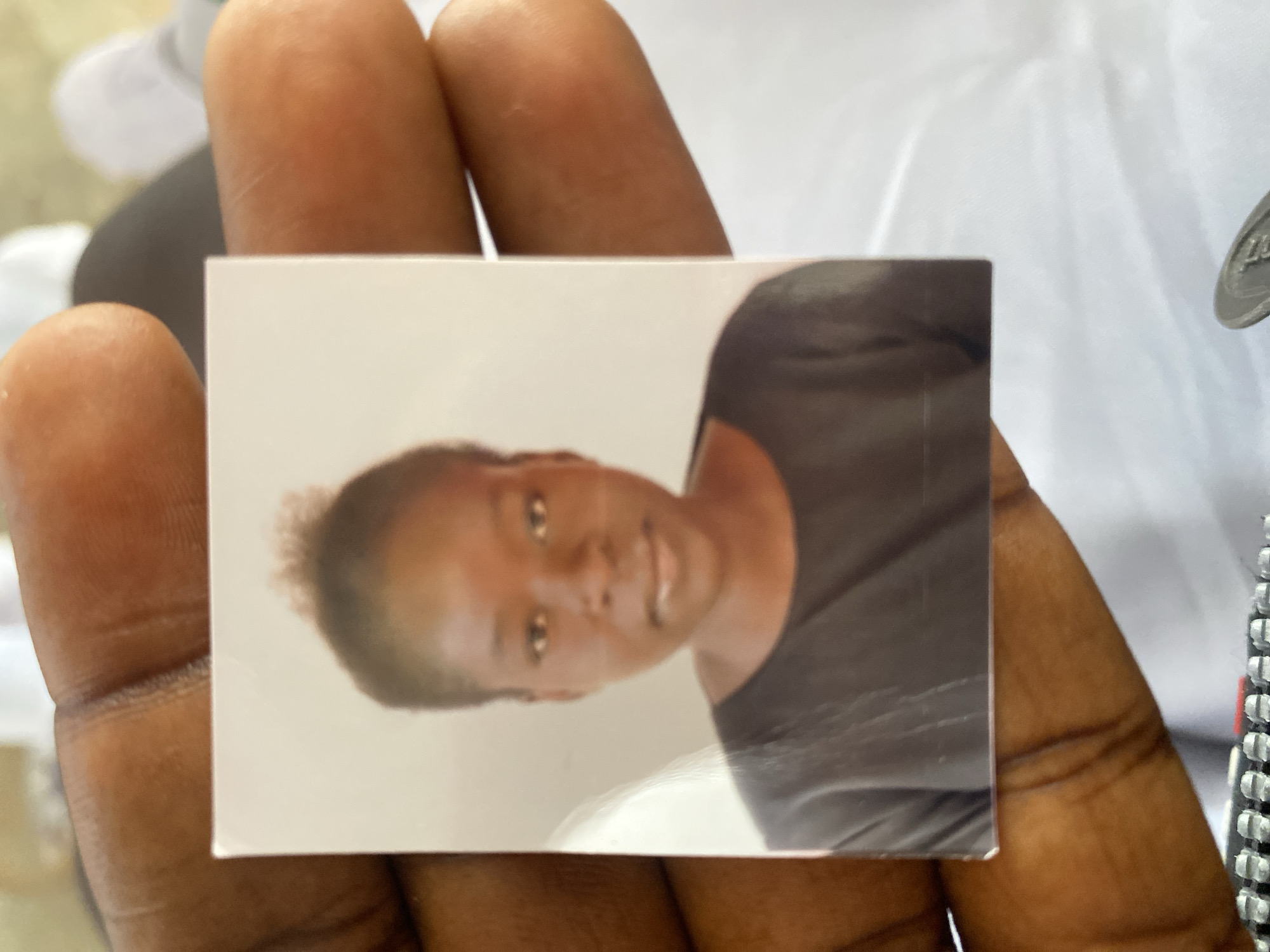 A headshot photograph of a young Black woman is held between the thumb and forefinger of a person's hand, obscuring the lower left and lower right corners of the photograph. The woman in the photograph has dark skin, short, dark, curly hair, and is wearing a dark crew-neck shirt. The background of the photograph is a plain, light color. The hand holding the photograph has dark skin and is resting on a white surface. The image was taken in Jimeta, Nigeria.