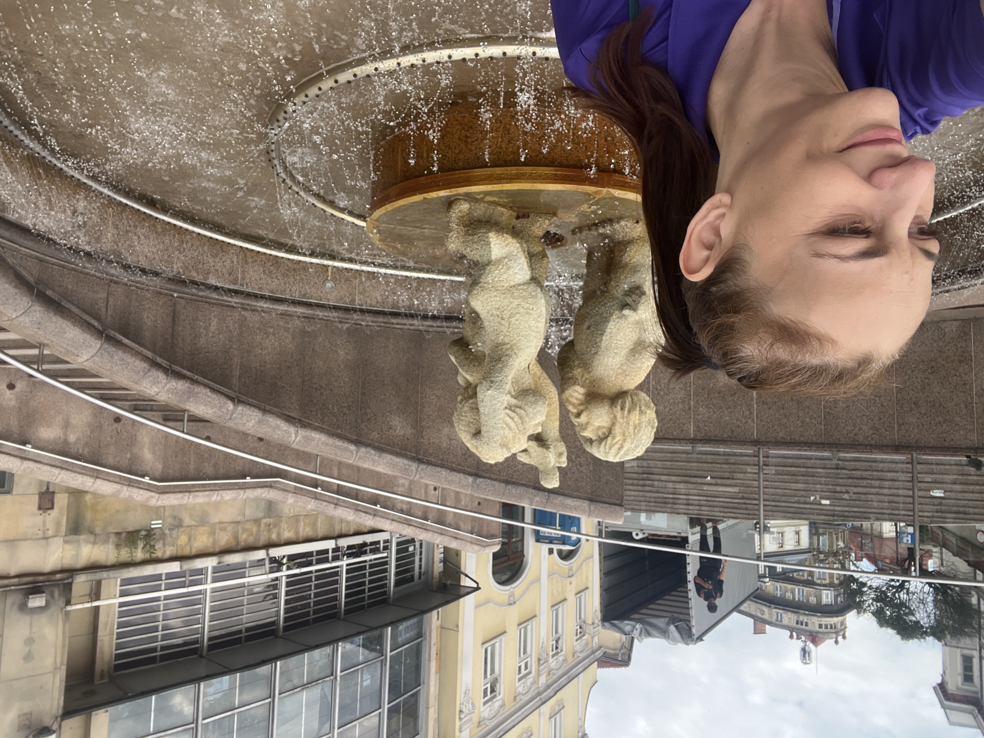In a public square in an unknown city, a woman is taking a selfie. She is wearing a purple shirt and has her hair tied back in a ponytail. In the background, there is a fountain with two stone statues of children. Behind the fountain, there are steps leading up to buildings and a truck with a tarp over its cargo. The sky is overcast.