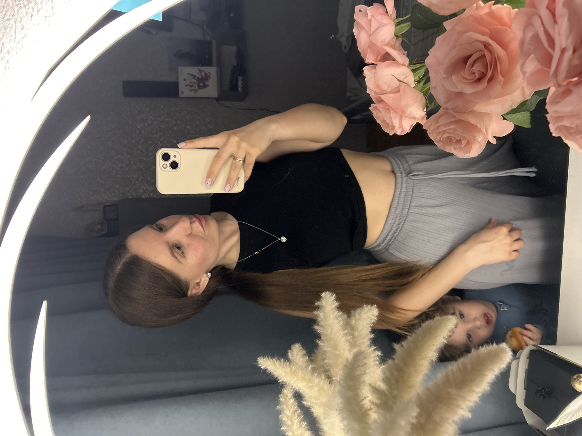 A woman takes a selfie in a mirror; in the reflection, a young child is visible. The woman is wearing a black top, gray pants, and a silver necklace. She is holding a smartphone. A bouquet of pink roses and beige pampas grass are in front of the mirror. The location is potentially Klin, Russia.