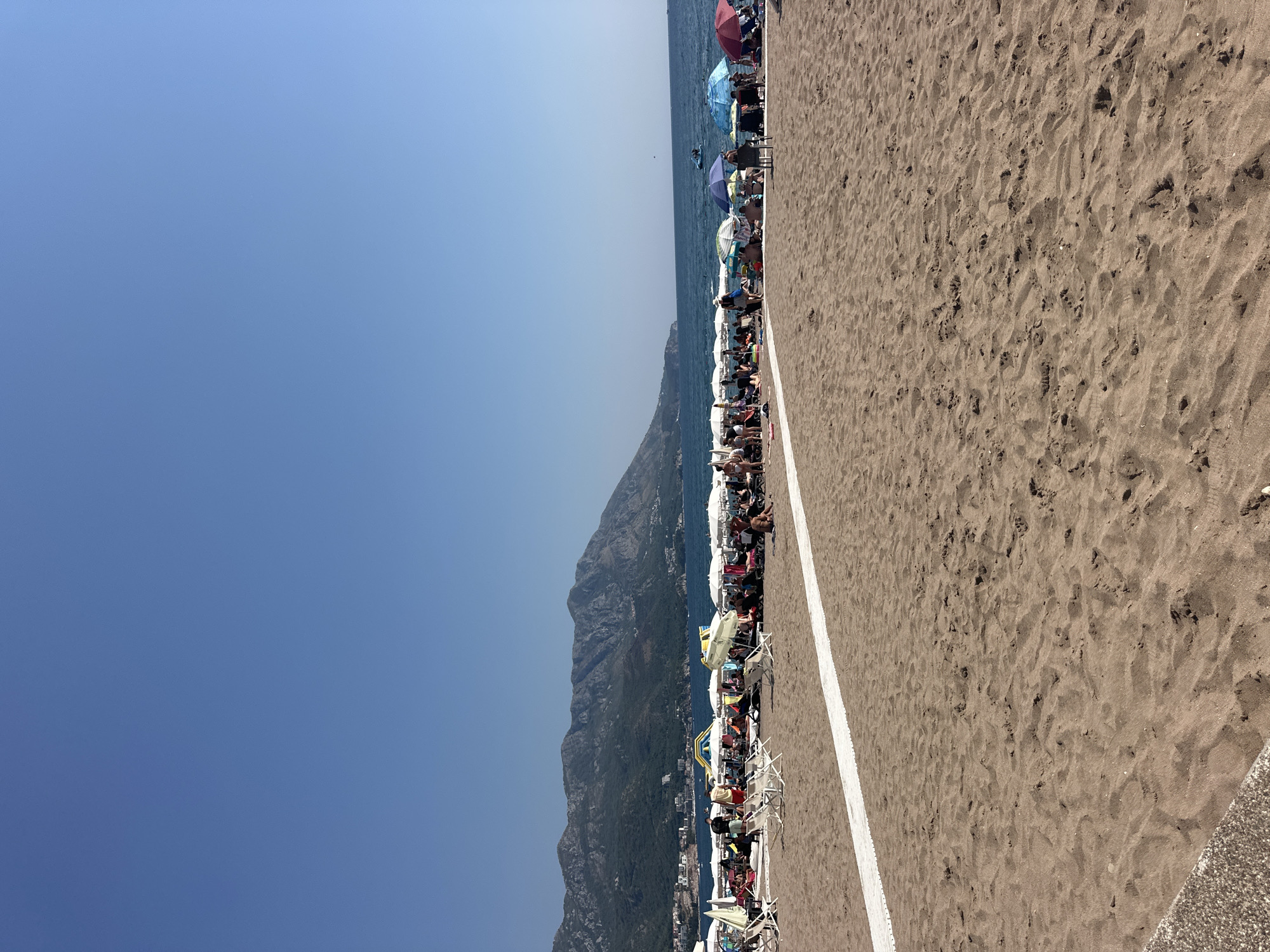 A wide shot captures a crowded beach on a sunny day, overlooking the Adriatic Sea and mountainous landscape in Boreti, Montenegro. The foreground is dominated by textured brown sand, with a white pathway laid out horizontally across the mid-ground. A densely populated area of beach umbrellas and loungers stretches along the shoreline, filled with numerous people sunbathing, sitting, and walking. The sea is calm with a few distant boats visible. The background features a city nestled against lush green mountains. The sky is a clear, bright blue.