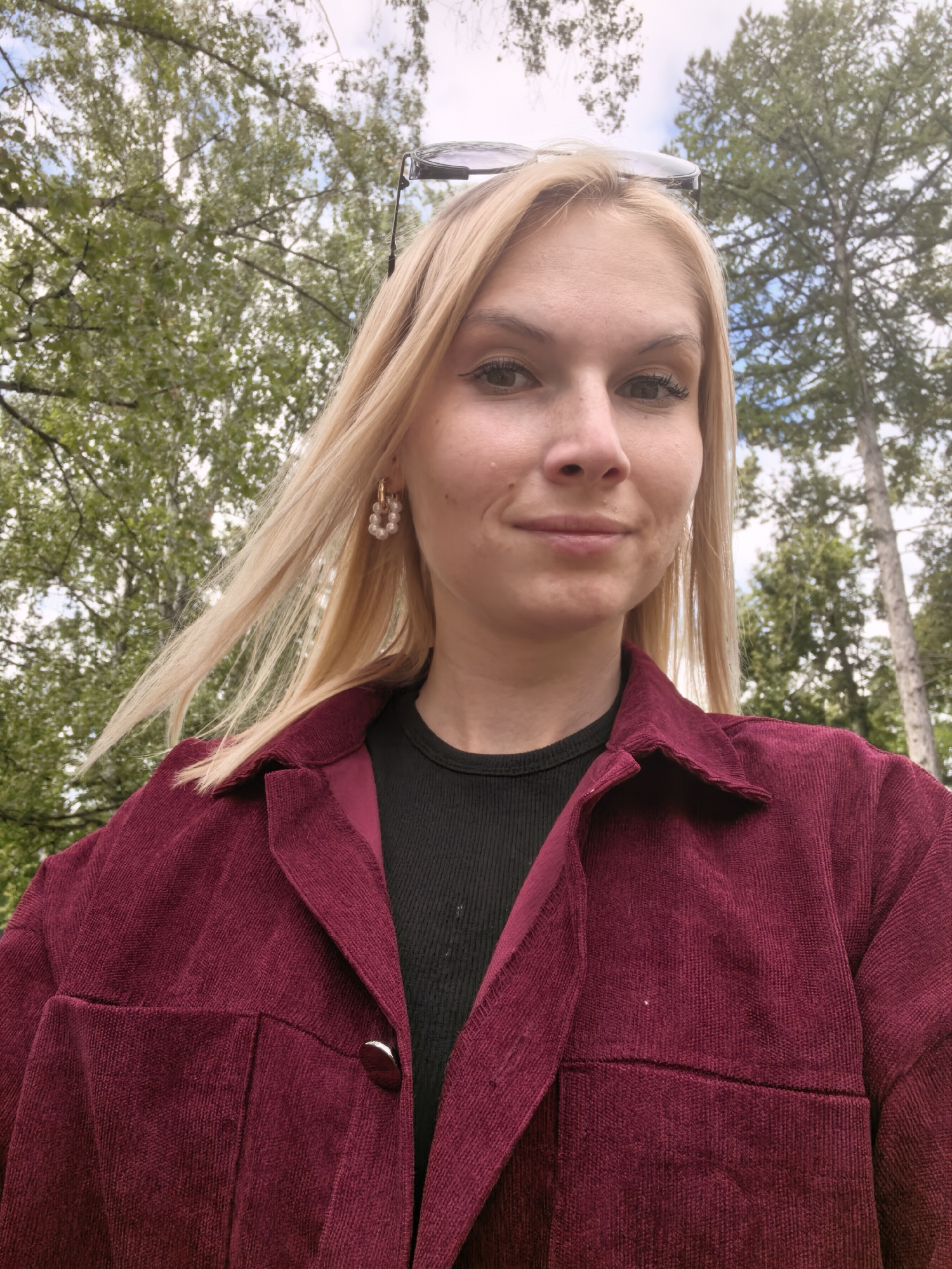 A close-up, selfie-style medium shot shows a young woman with blonde hair and fair skin outdoors. She is wearing a dark ribbed shirt under a dark red corduroy shirt jacket. Her hair is blowing slightly to her left, and she is wearing pearl earrings. Sunglasses are perched on top of her head. The background consists of green trees and a light sky.