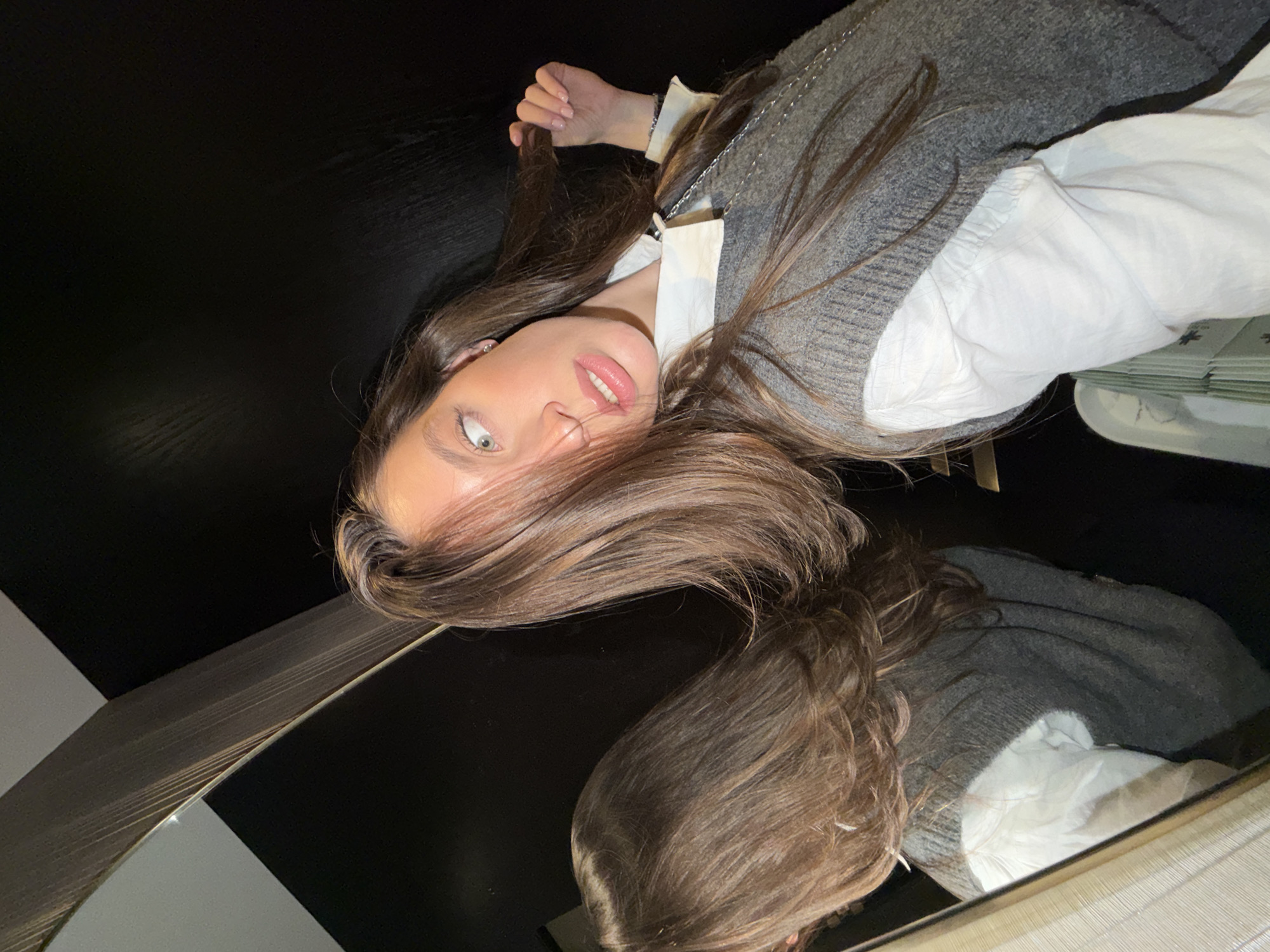 A young woman is lying on a shiny black surface, possibly a table or countertop, with her head tilted towards the viewer. The background is a combination of black and a reflection showing a gray surface. She has long brown hair, a fair complexion, and green eyes. She's wearing a white collared shirt under a gray sweater. The image appears to be taken at an unusual angle, perhaps as a selfie or from a low vantage point. There's no explicit evidence indicating the location is Budva, Montenegro.